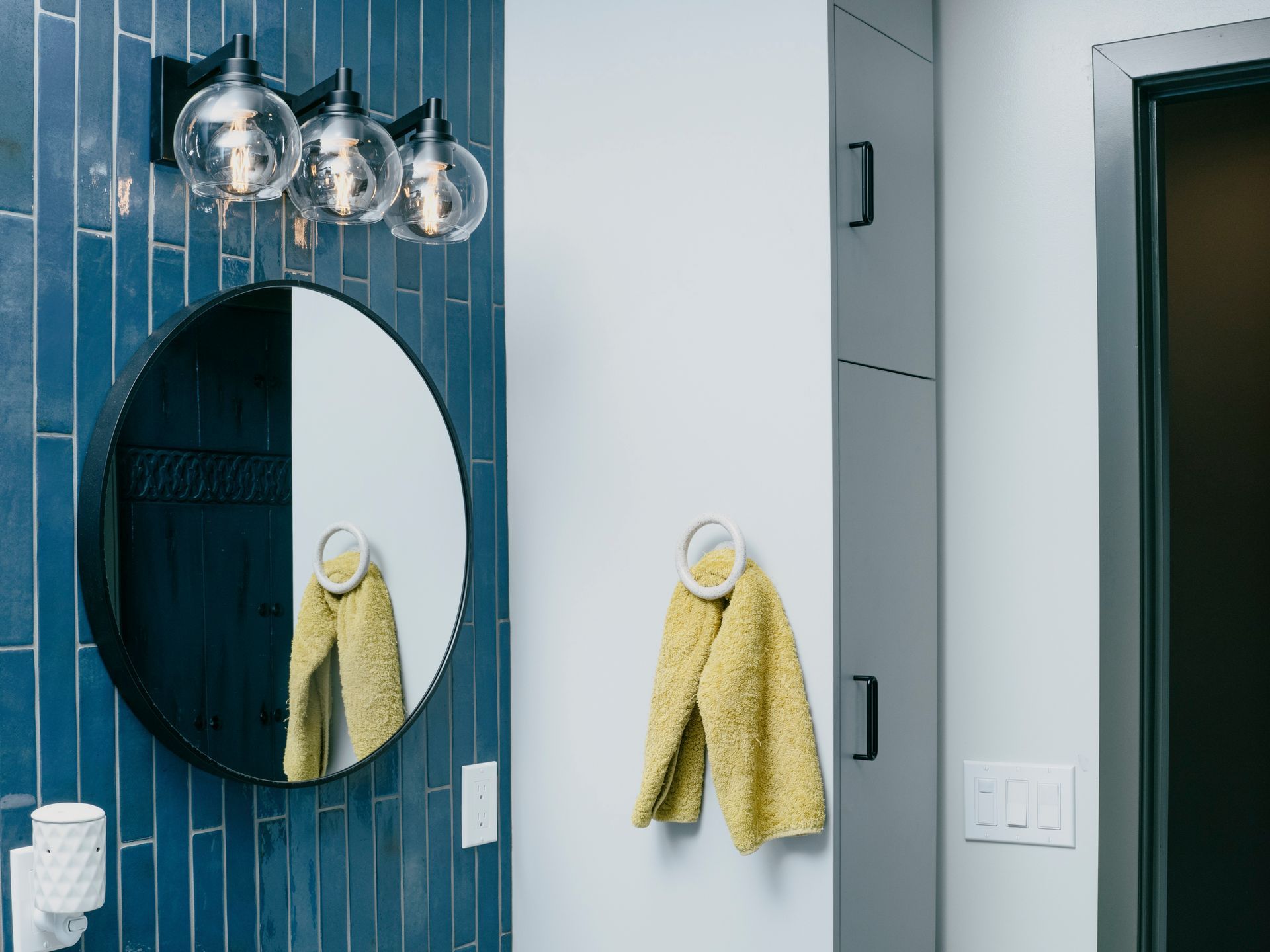Bathroom with blue tiled wall, round mirror, globe light fixture, and yellow towel.