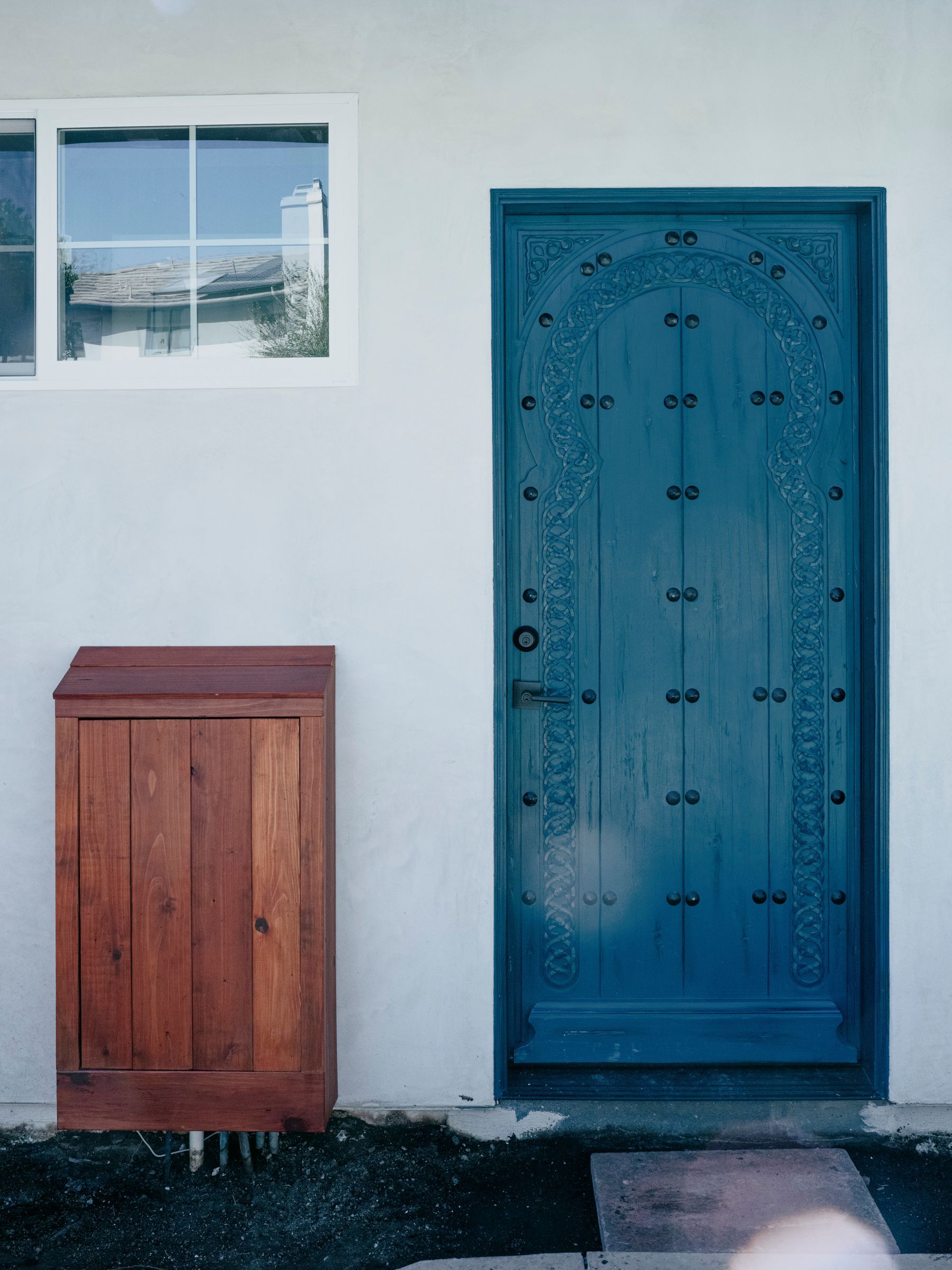 Blue ornate door next to a small window and a wooden mailbox on a white wall.