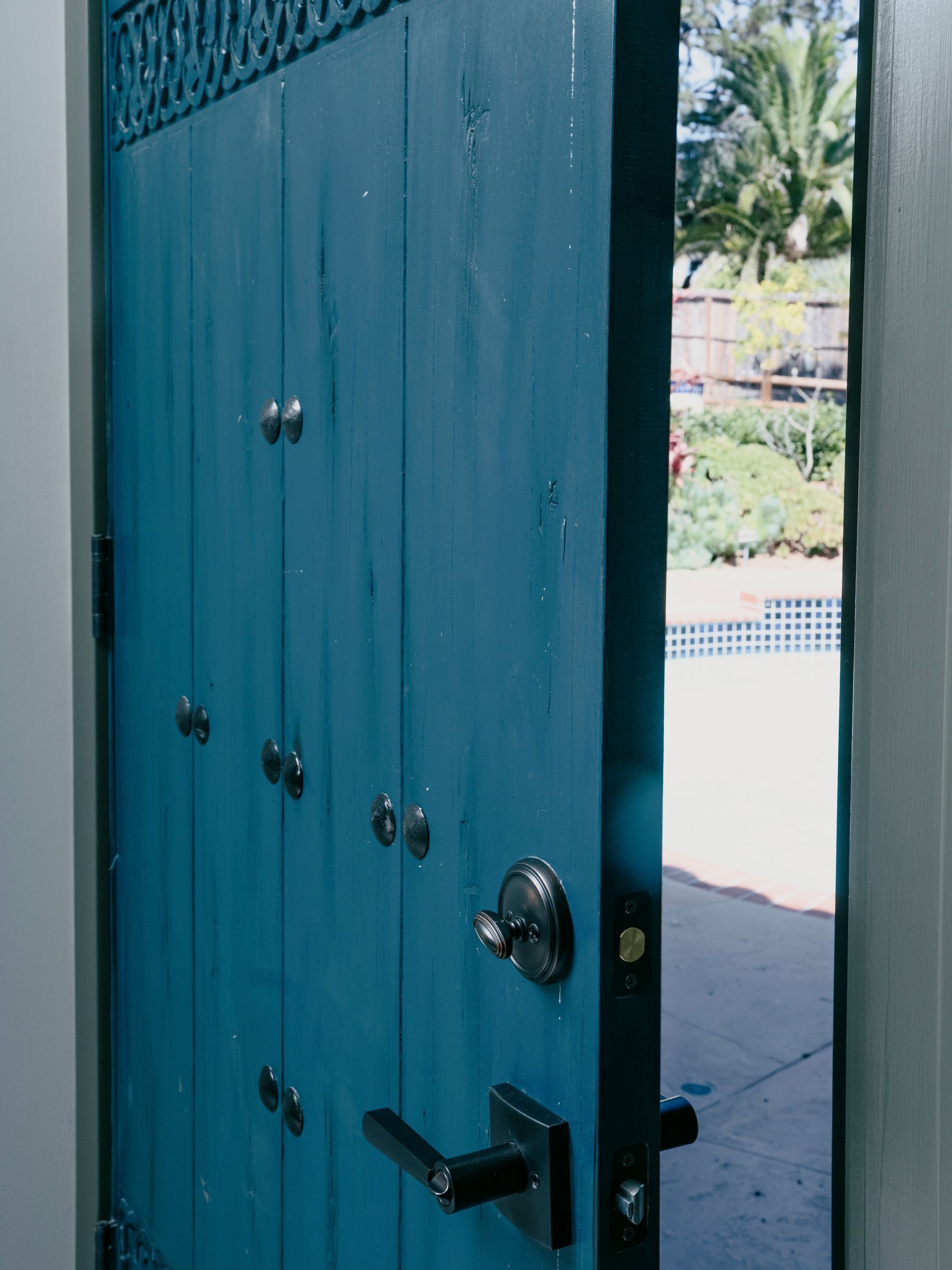 Blue wooden door with decorative studs, ajar, revealing a pool and garden.