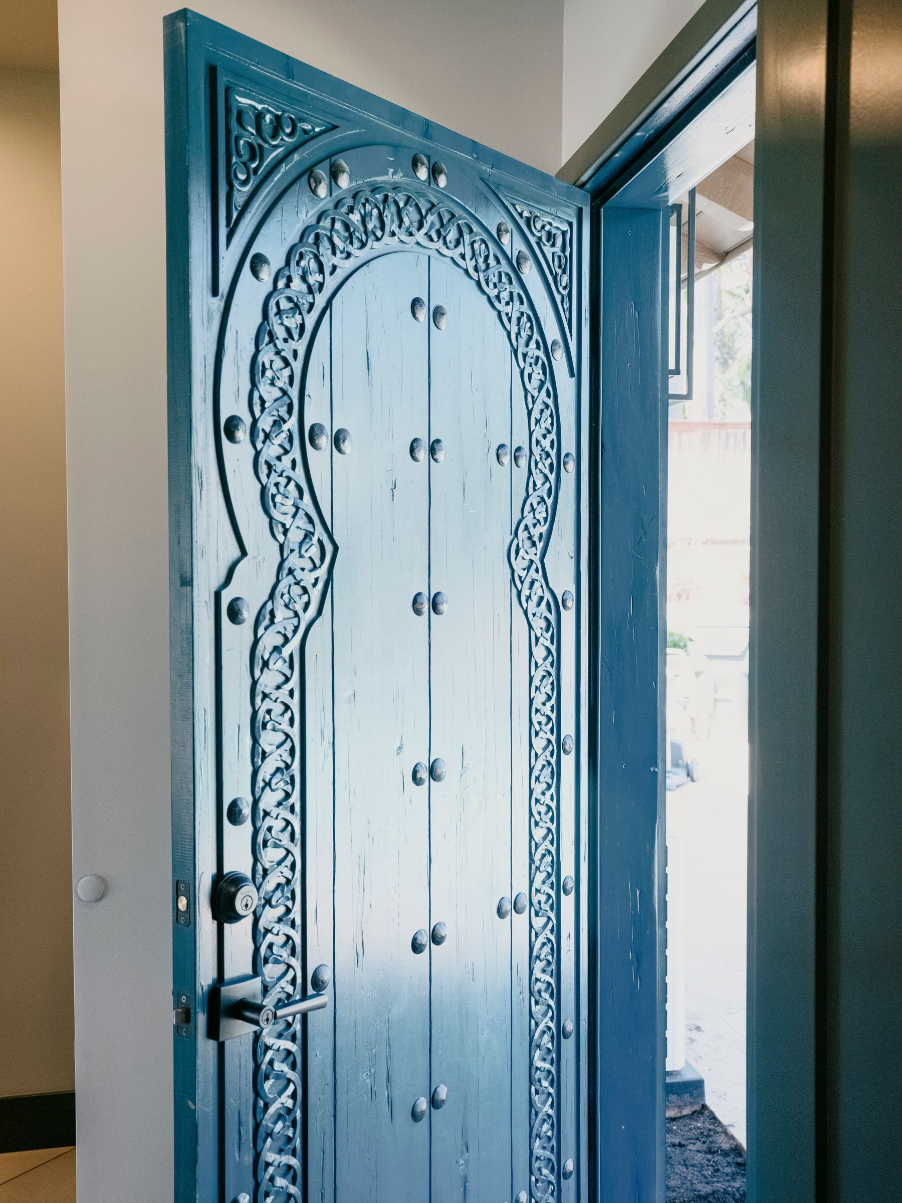 Blue, arched doorway with ornate carvings and small studs. Opens into a bright space, likely an entryway.