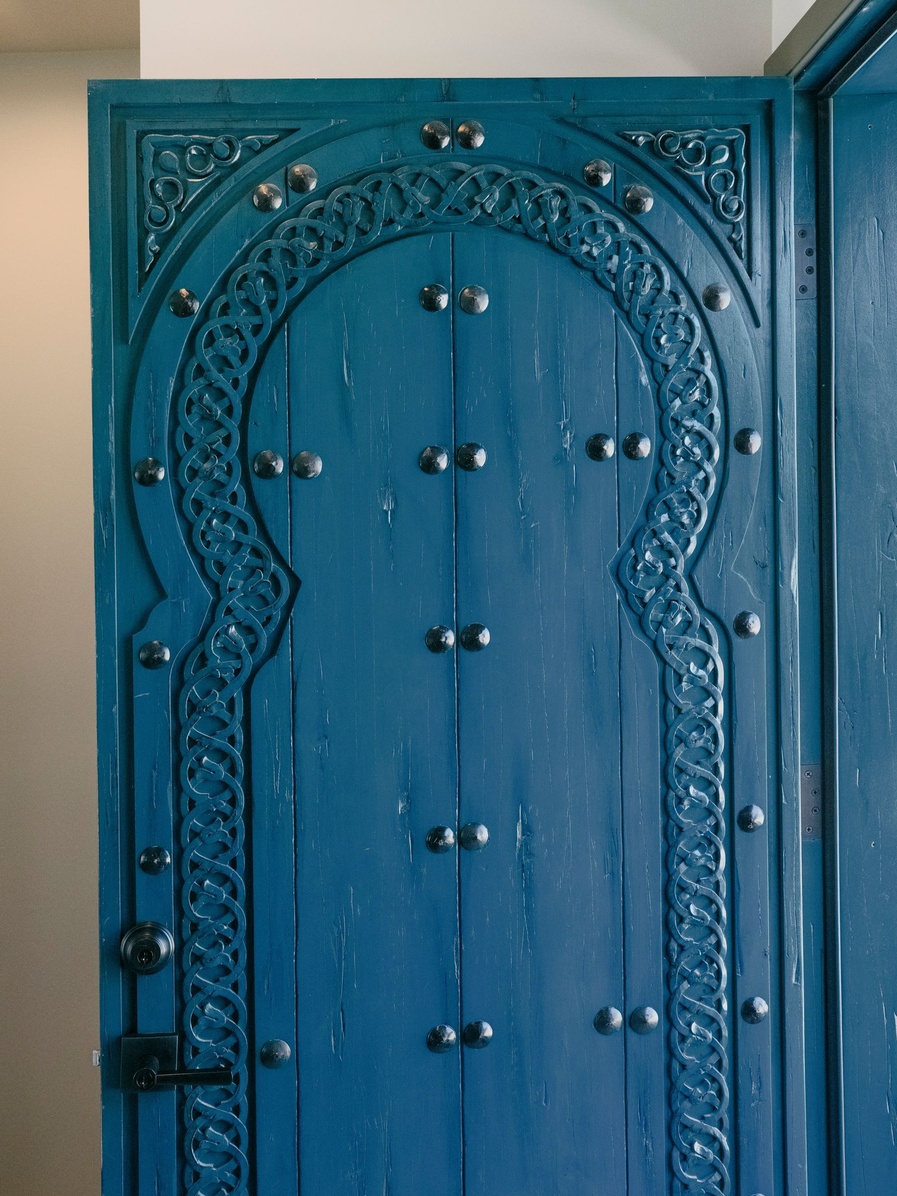 Blue, arched wooden door with ornate carvings and metal studs, in a bright setting.