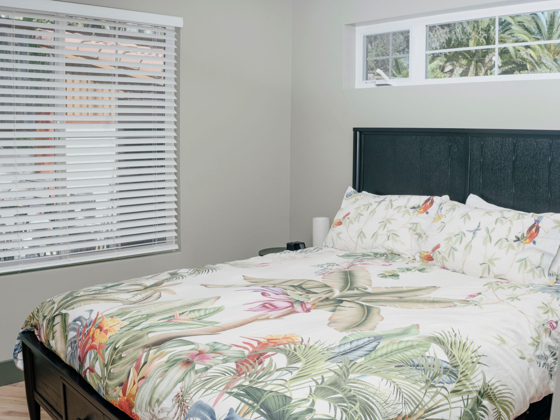 Bedroom with bed, floral bedding, window with blinds, and a small window above the headboard.
