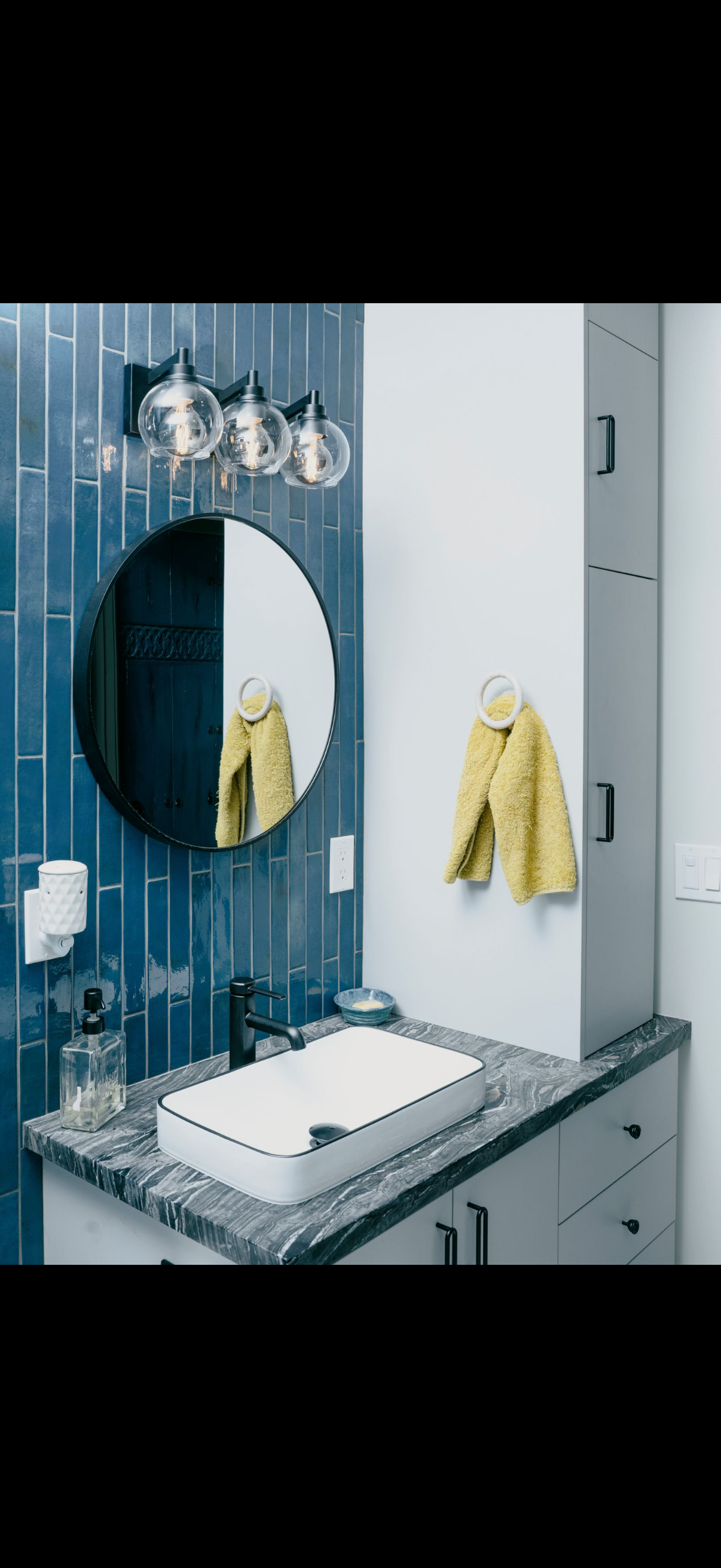 Modern bathroom with blue tile, a round mirror, and a white sink. A yellow towel hangs.