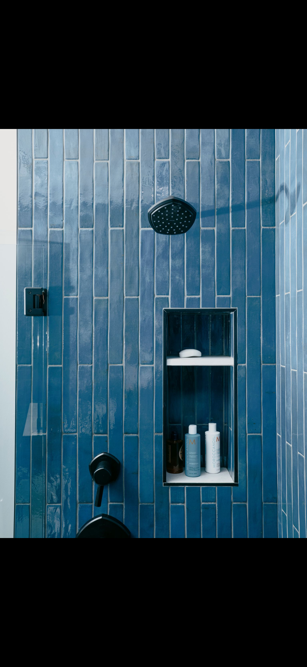 Blue tiled shower with black fixtures and built-in shelf holding toiletries.