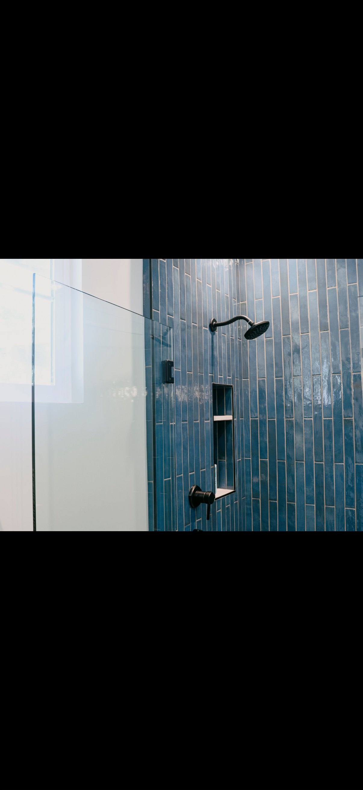 Shower with a dark shower head and fixtures against a textured blue wall, behind a glass panel.