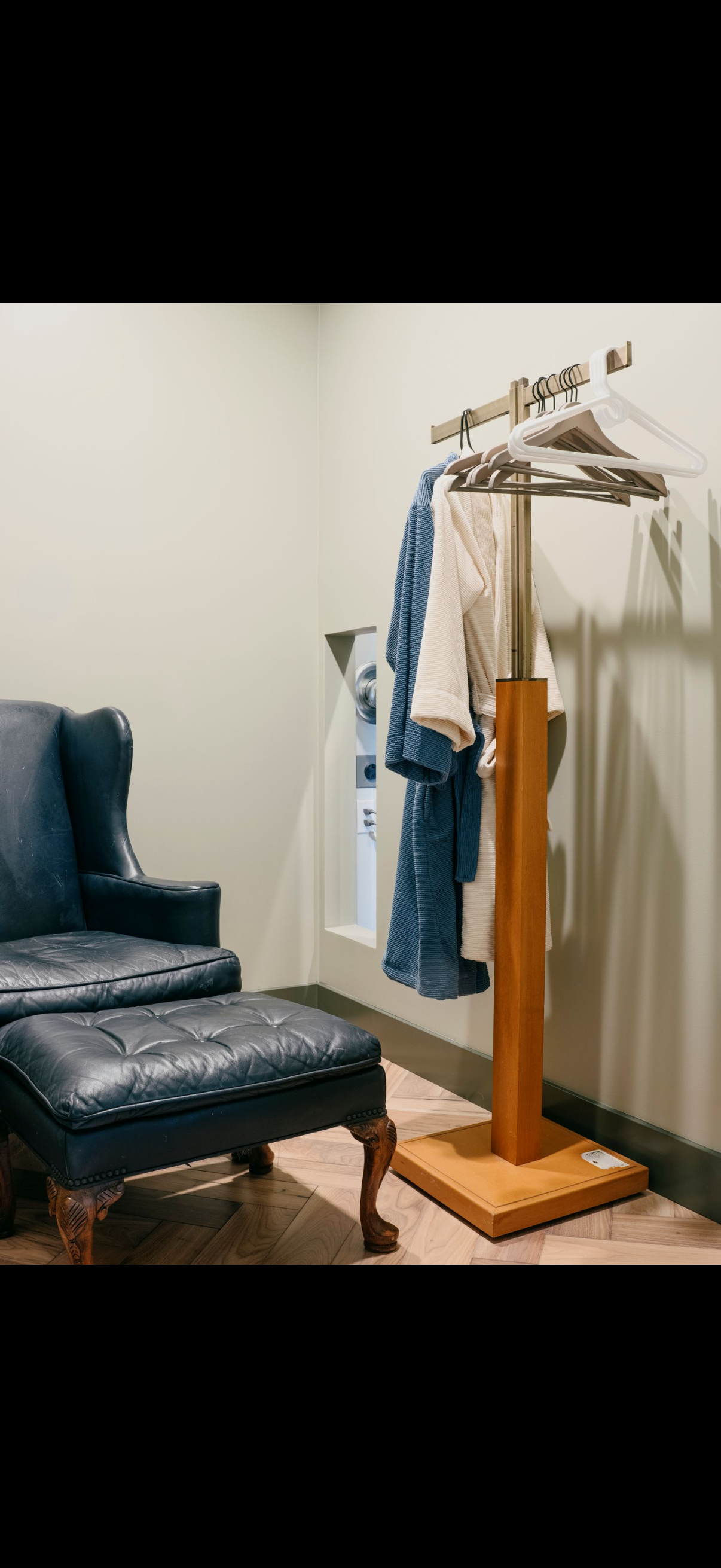 A room with a blue leather chair and ottoman, and a wooden clothes rack holding garments.