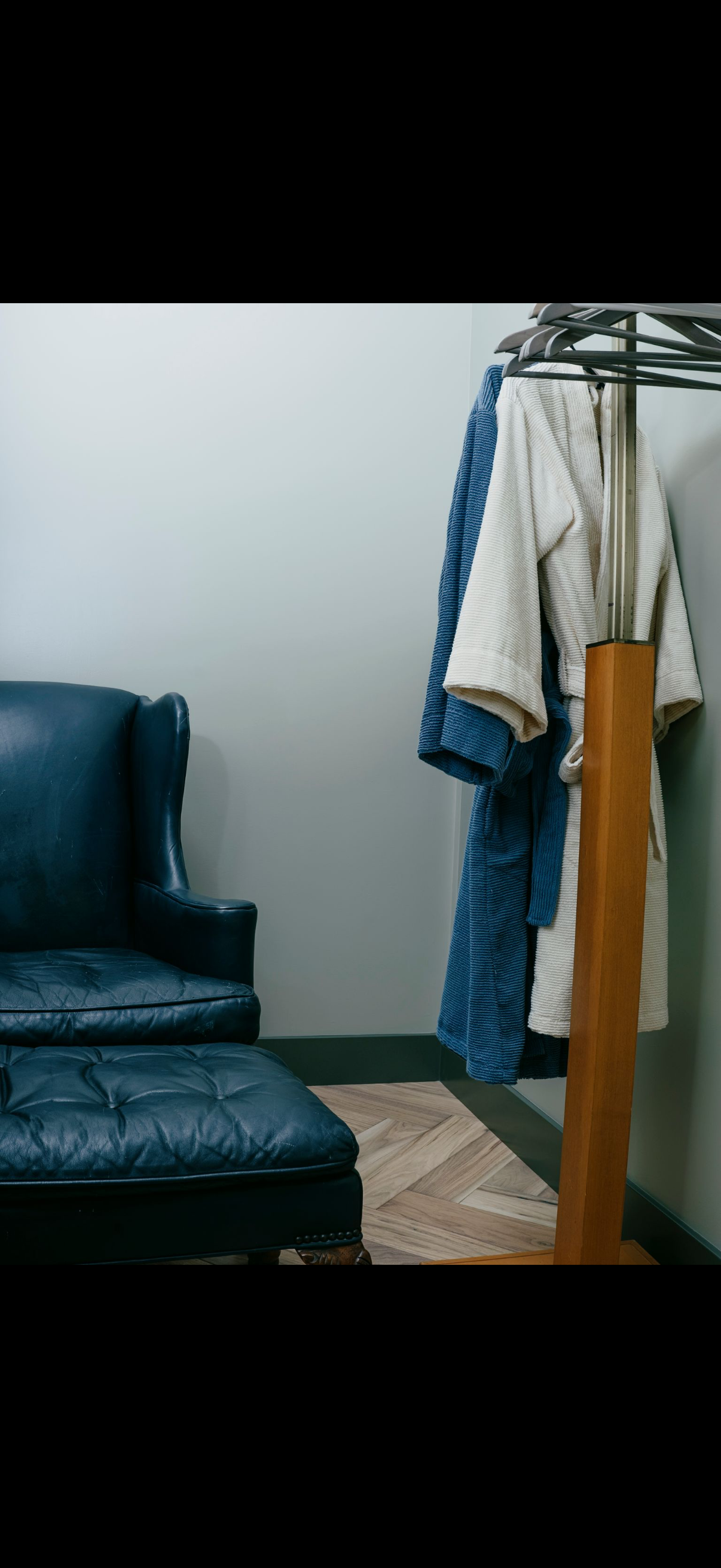 A blue leather armchair with an ottoman, two bathrobes on a rack, in a room.