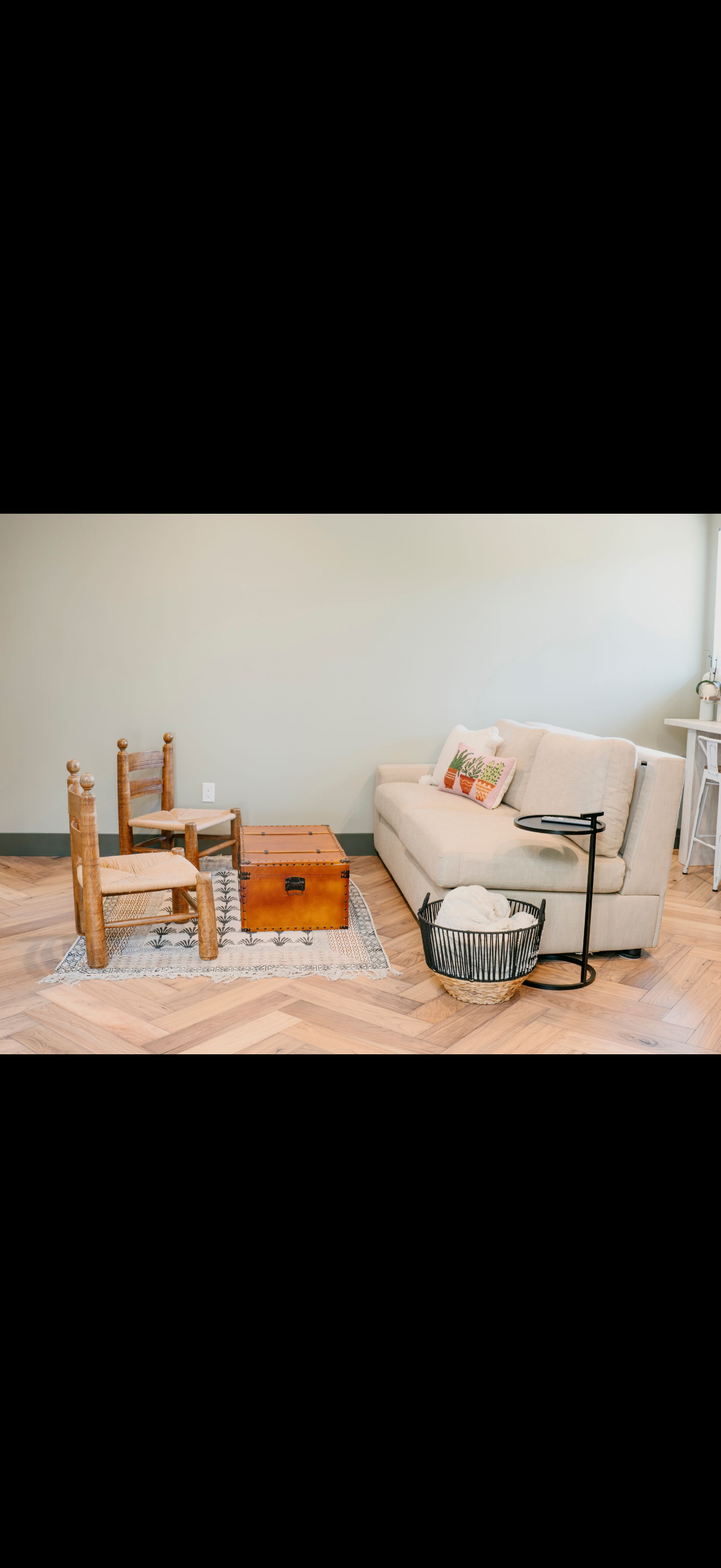 A living room setting with a white sofa, two wooden chairs, a chest, and patterned rug.