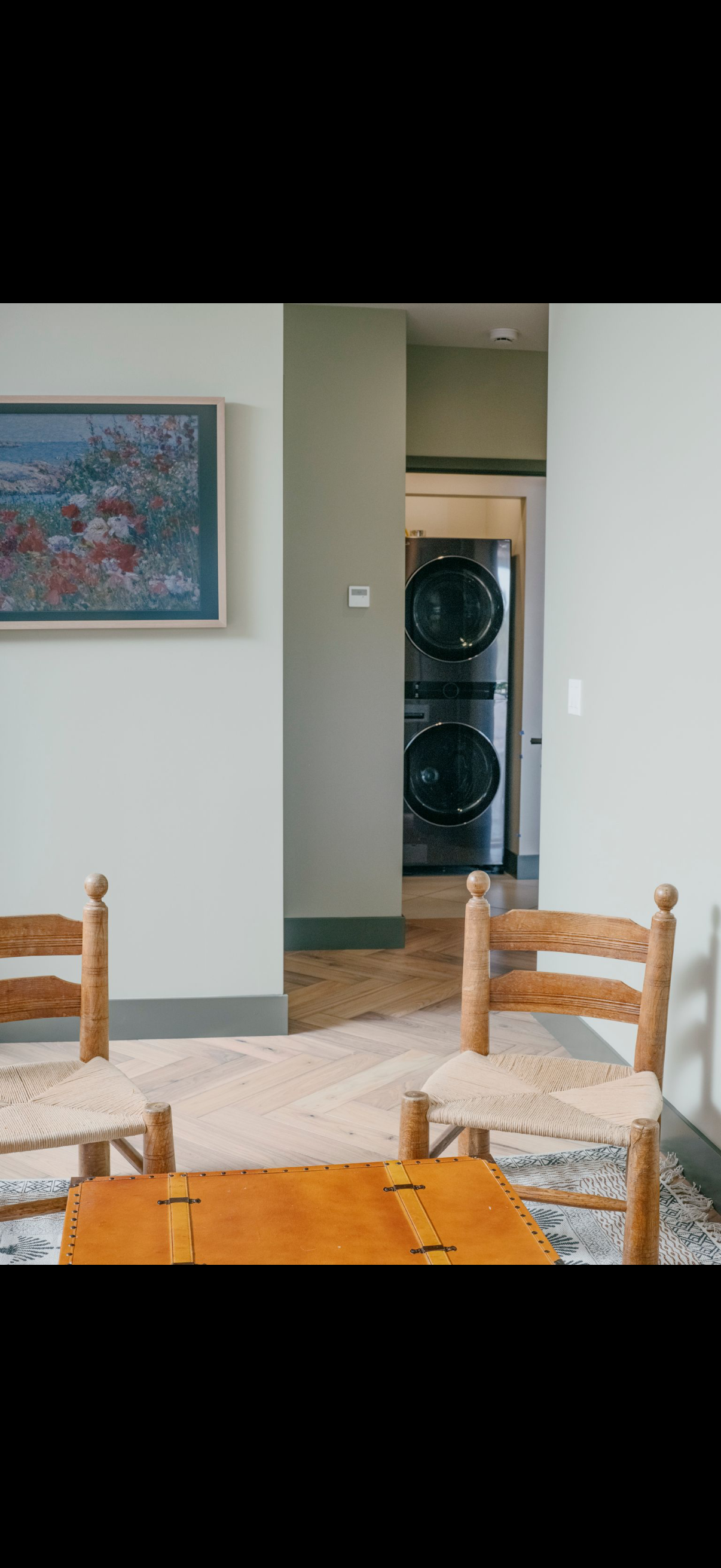 Two wooden chairs with woven seats flank a small table in a room. A laundry room is visible in the background.