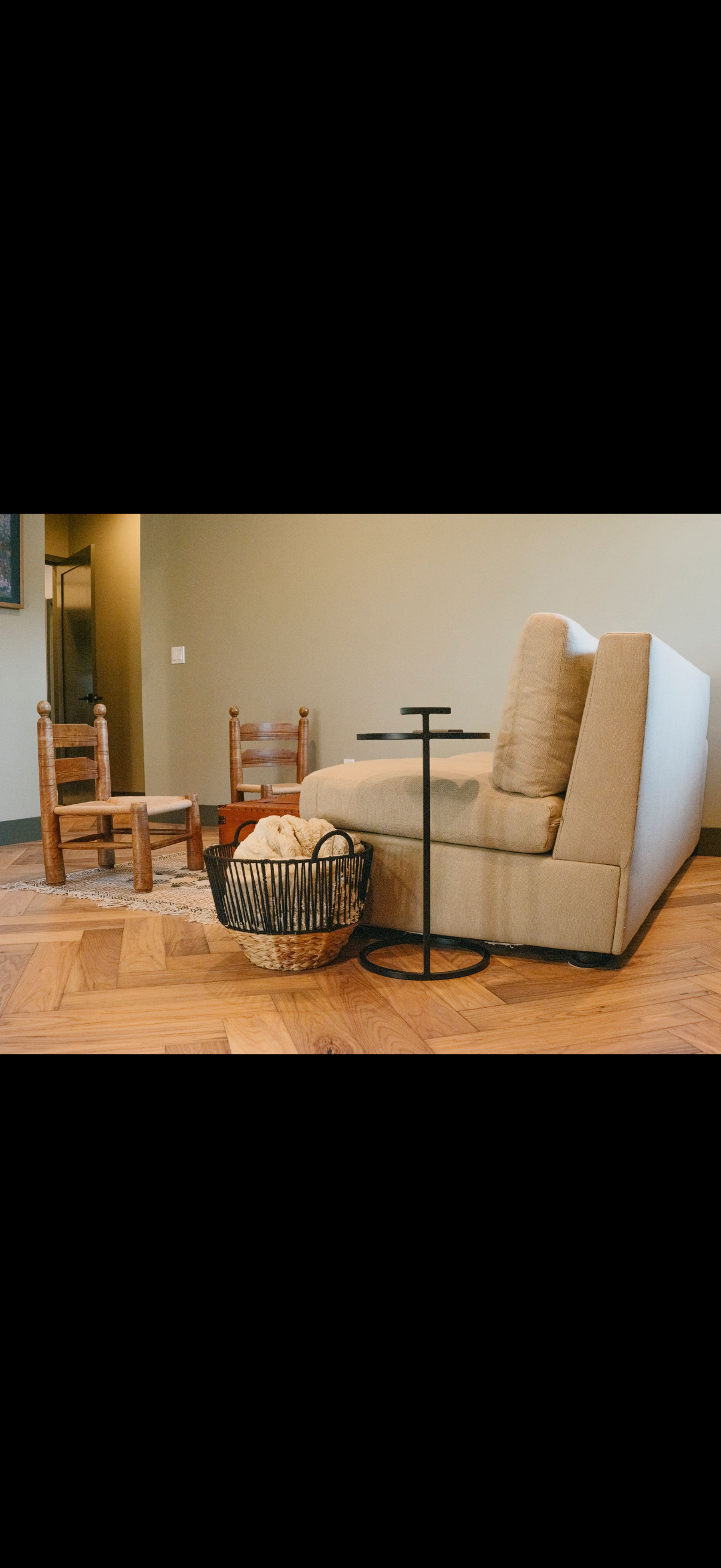 A living room with a couch, small table, basket, and chairs. Light-colored walls and wood floors.