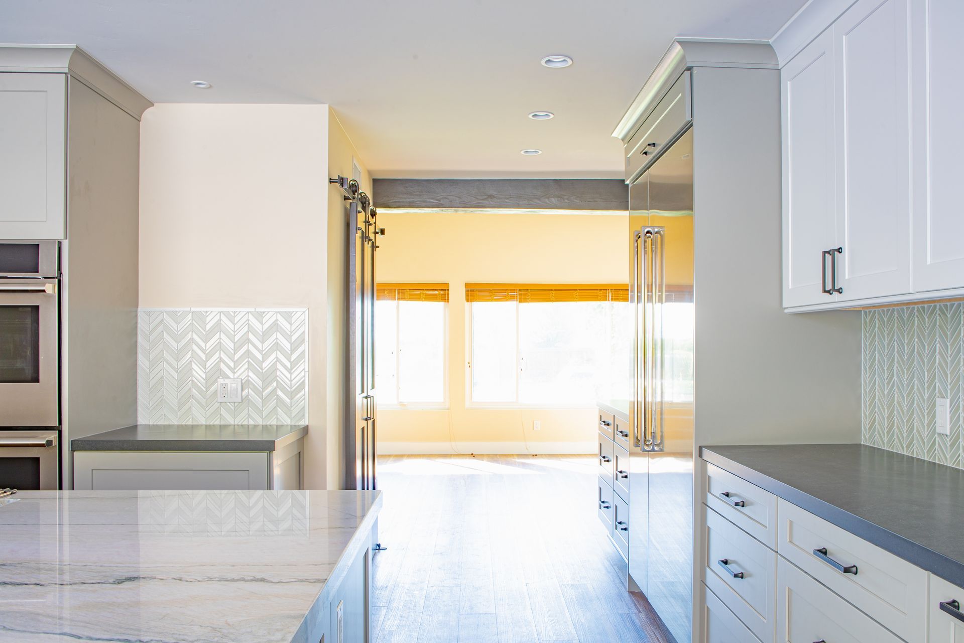 Bright kitchen interior with white cabinets, gray counters, and a doorway leading to a window-lit room.