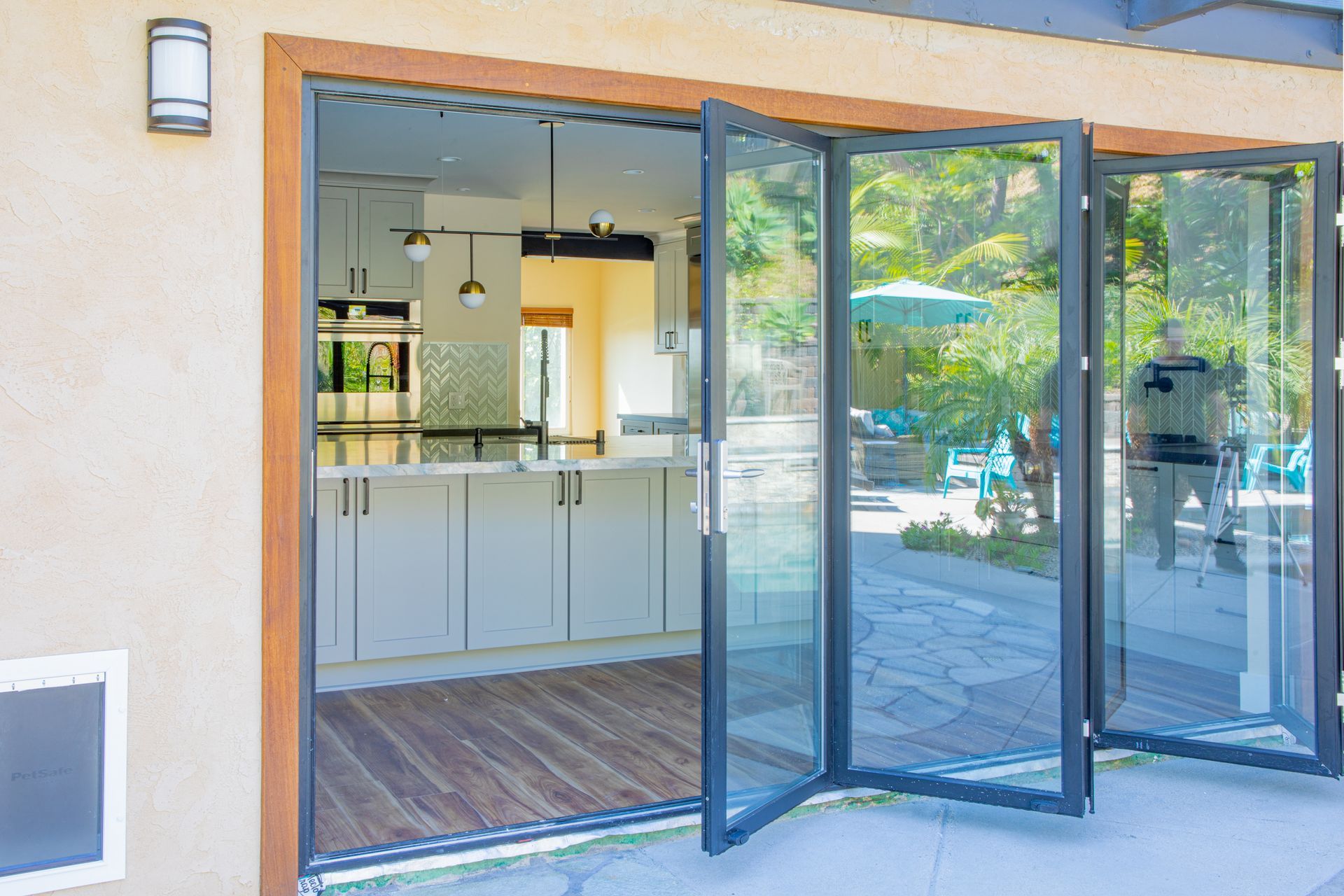 Open bifold doors reveal kitchen, patio. Gray doors, light cabinets, wood floor. Person reflected in glass.