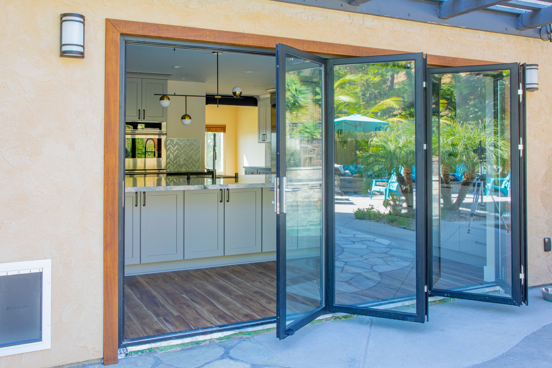 Folding glass doors open to a kitchen, connecting to a patio with outdoor seating.