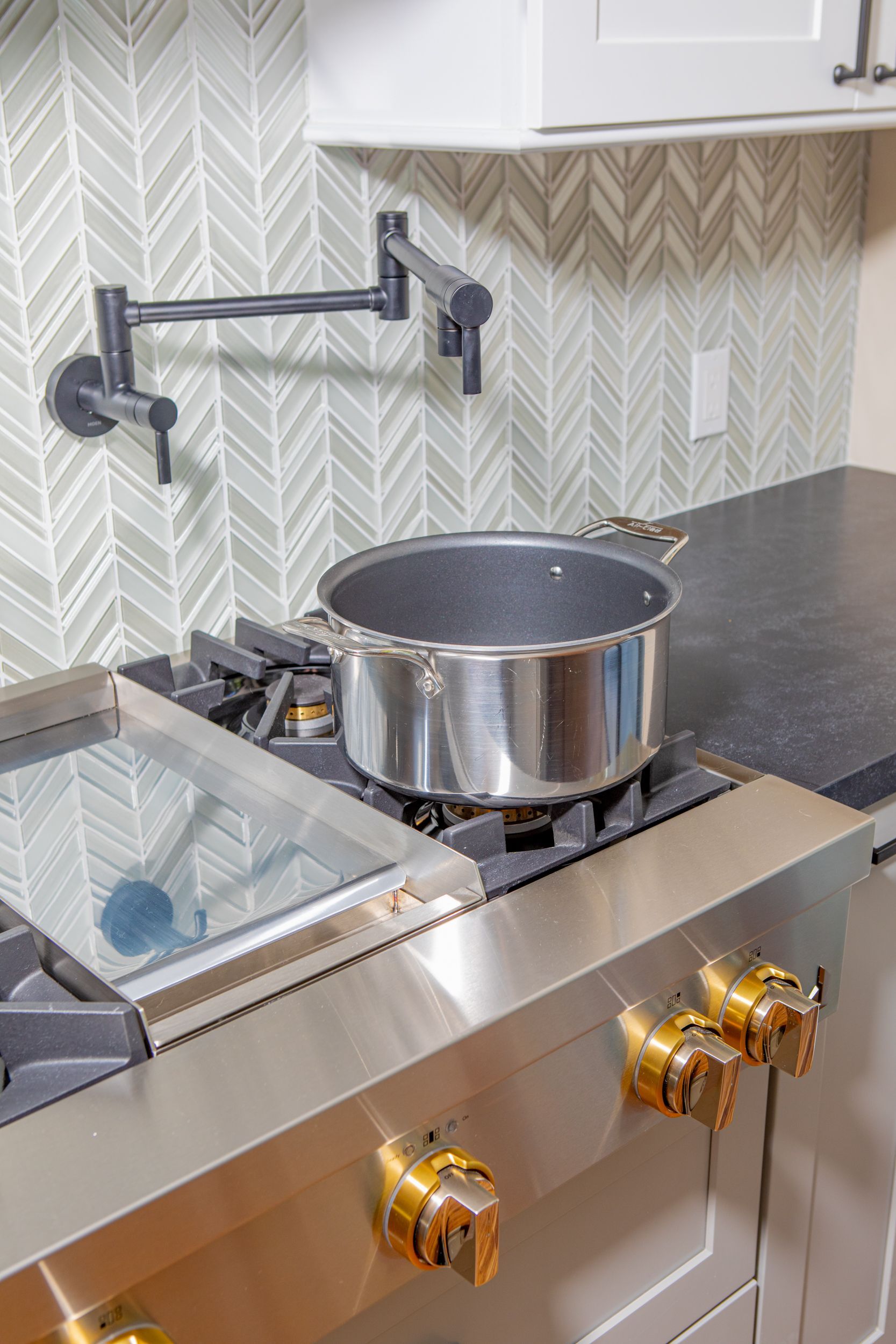 Stainless steel pot on a stovetop with a wall-mounted pot filler against a gray herringbone tile backsplash.