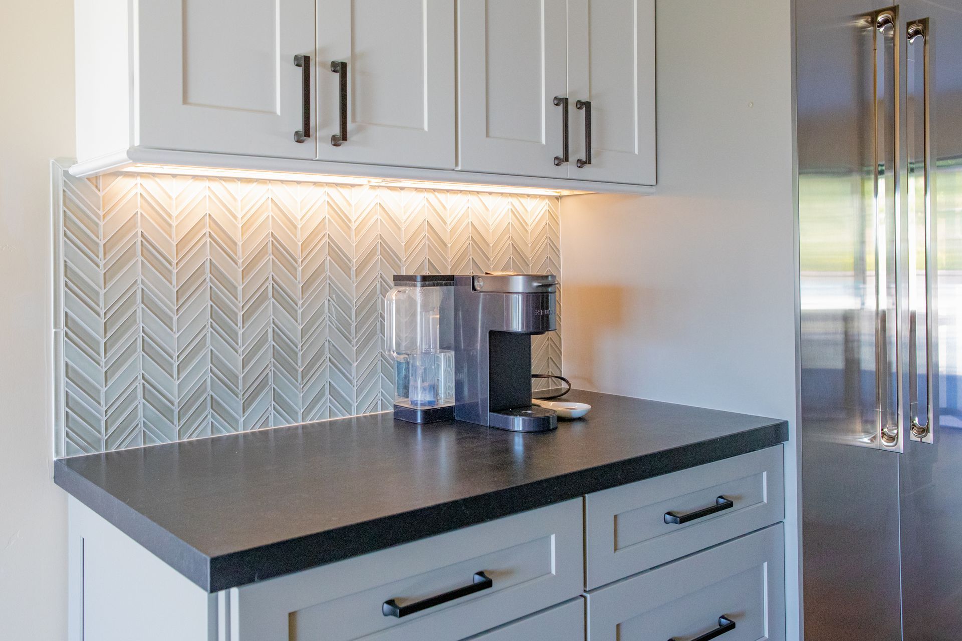 Kitchen coffee station with light cabinets, gray countertop, herringbone backsplash, and Keurig.