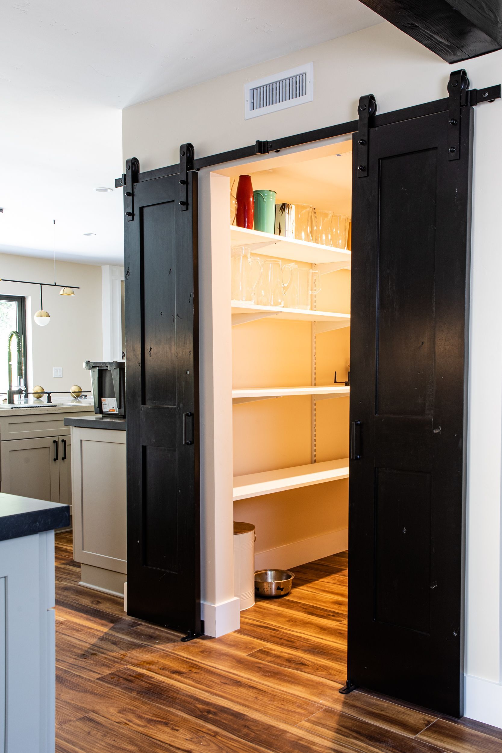 Black sliding barn doors open to reveal a pantry with white shelves; warm lighting and jars are visible.