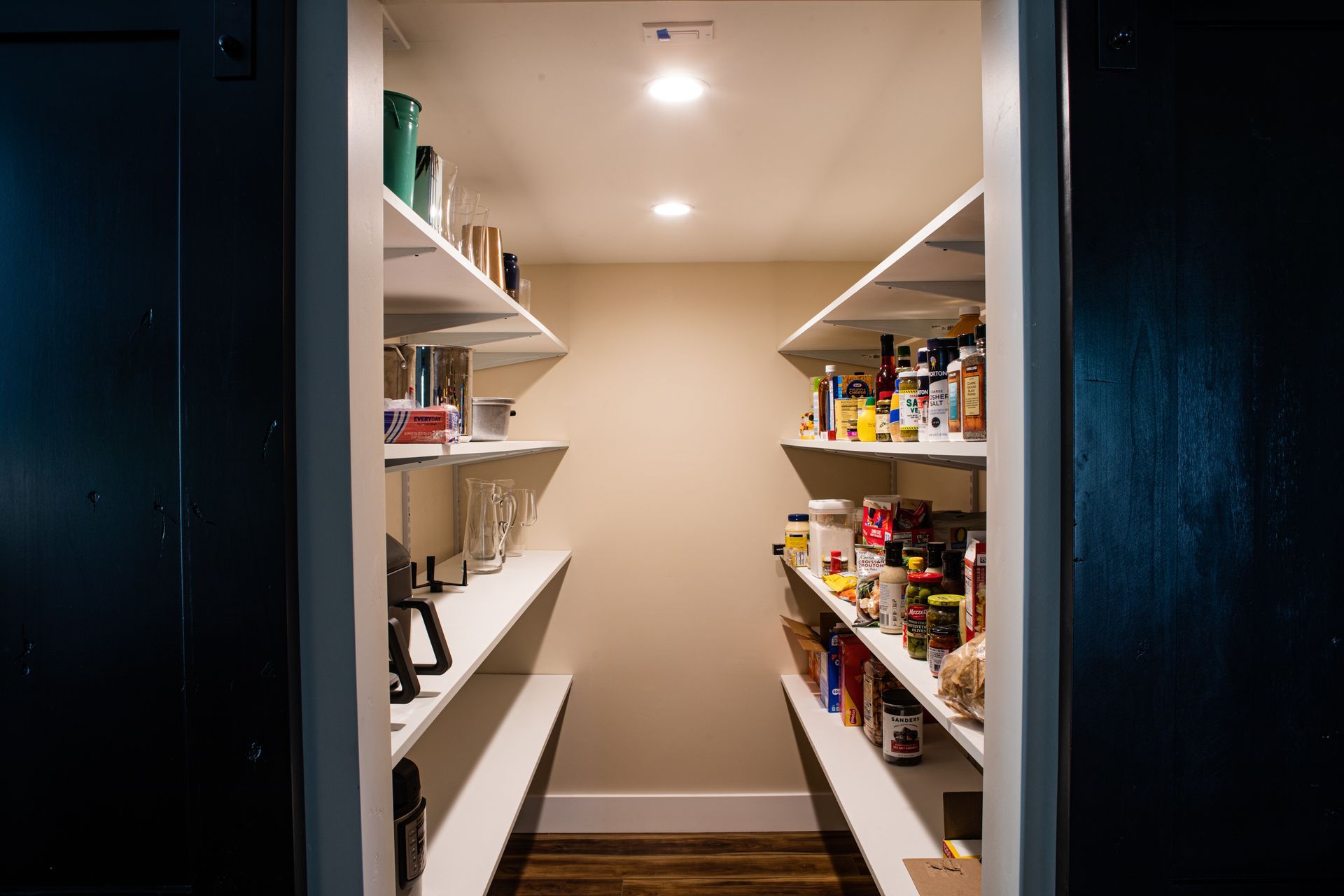 Pantry with white shelves on both sides, filled with food items. Beige walls, recessed lighting, and dark door frames.