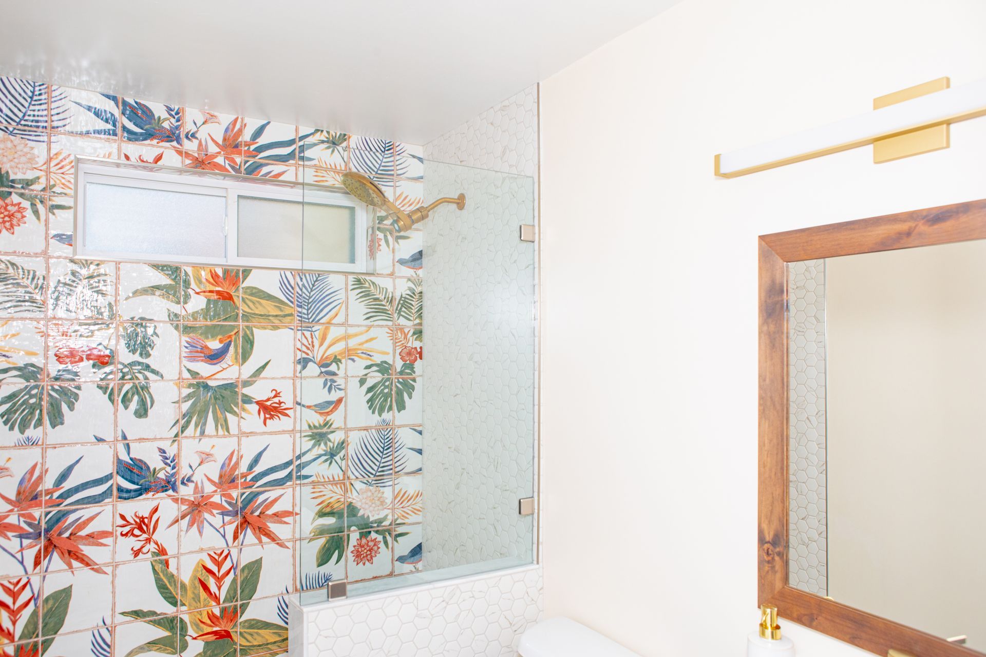Bathroom with floral tile shower, glass door, wood-framed mirror, and gold light fixture.