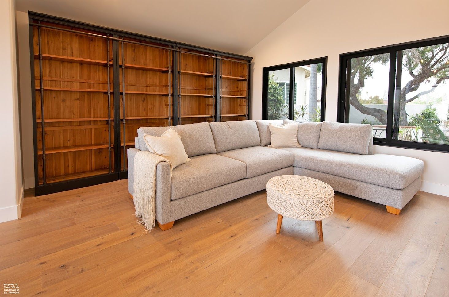 Living room with built-in wooden bookshelves, gray sectional sofa, and an ottoman, with large windows.
