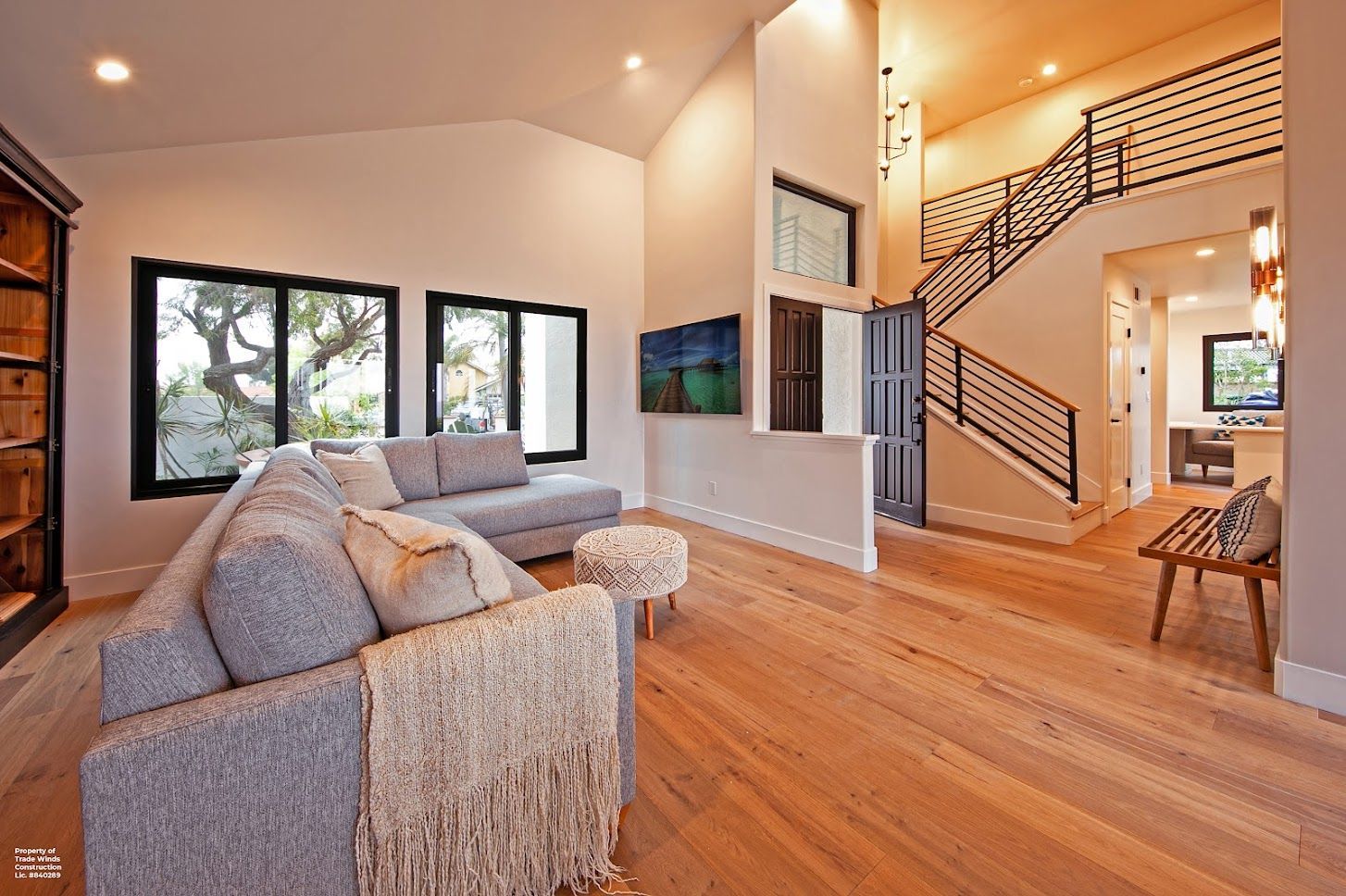 Modern living room with grey sofa, hardwood floors, black-framed windows, and a staircase.