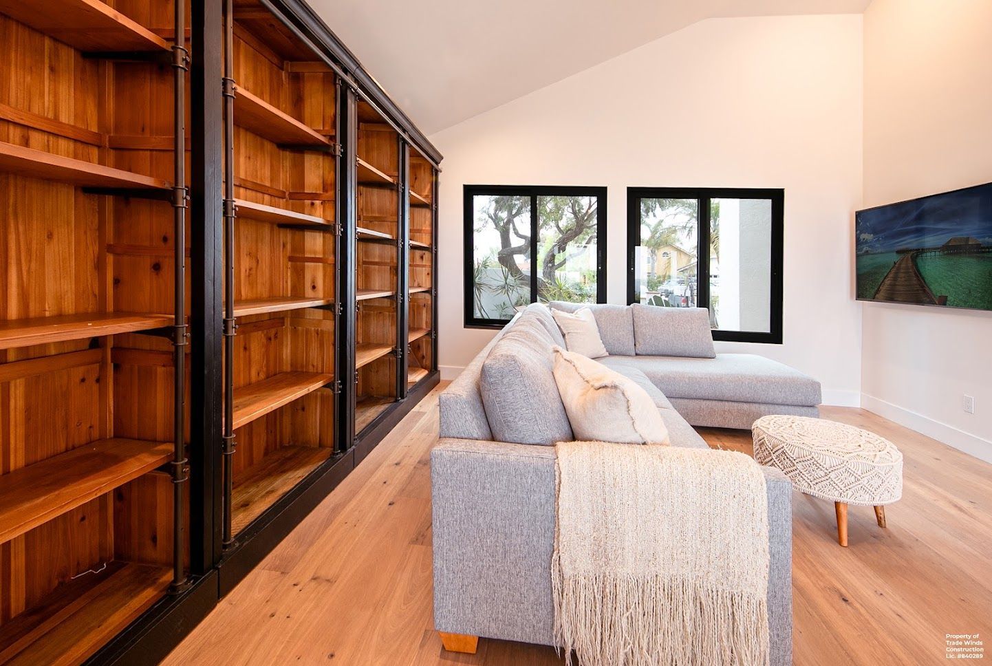 Wooden bookshelves line a light-filled living room with a gray sectional, ottoman, and artwork.
