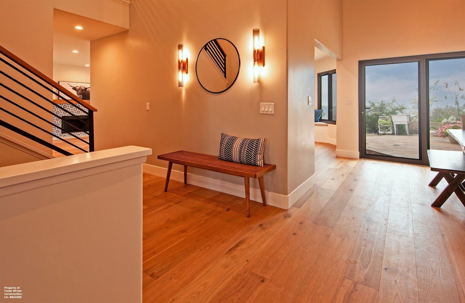 Hallway with wooden floor, bench with pillows, artwork, and wall sconces; open to an outdoor patio.