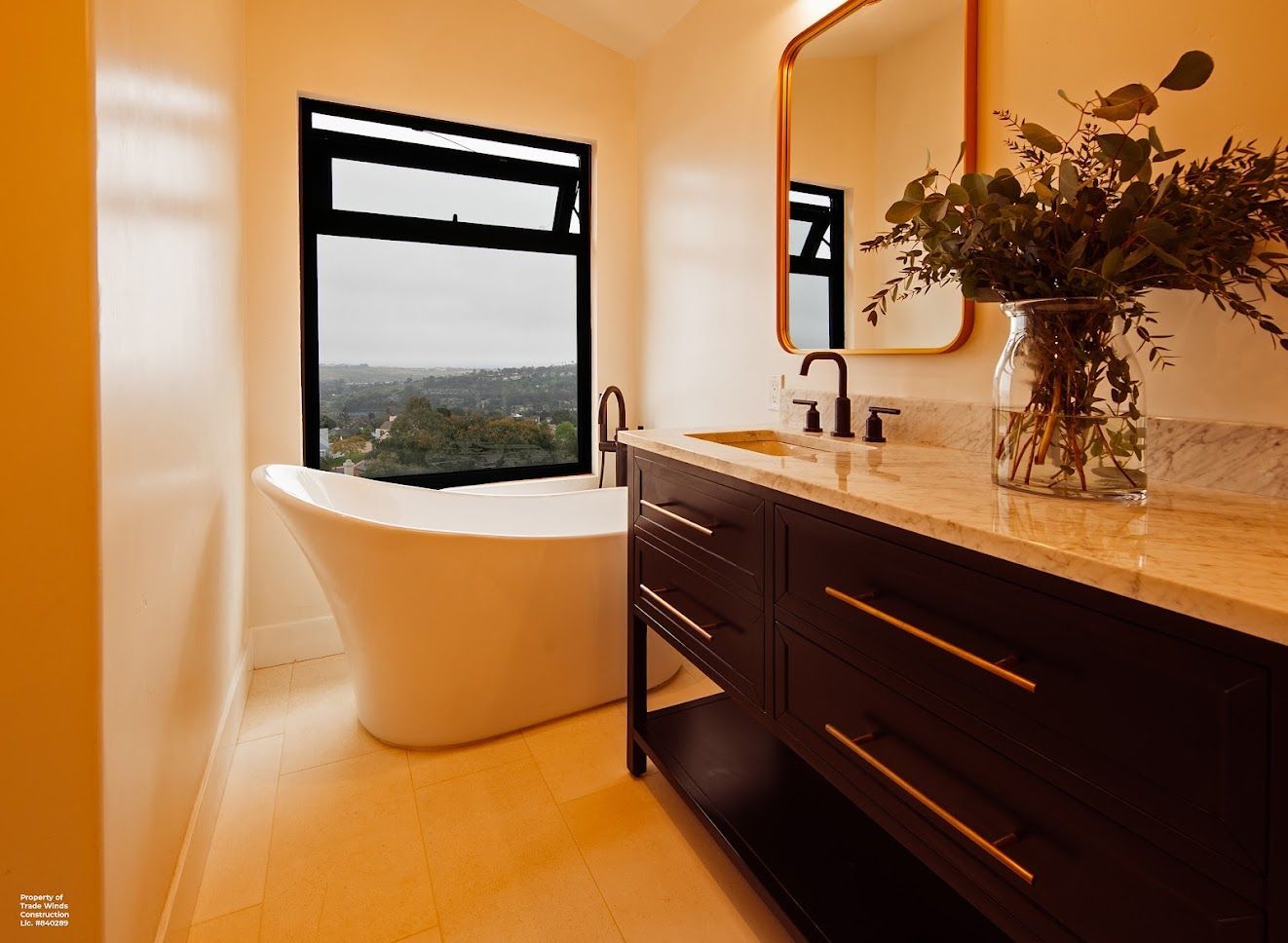 Bathroom with a white tub near a window with a view, dark brown vanity, gold-framed mirror, and floral arrangement.