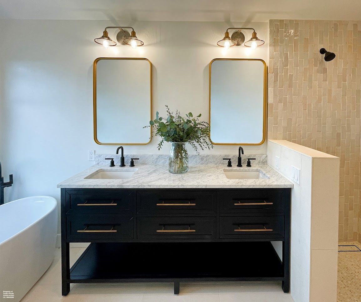 Bathroom with black vanity, gold-framed mirrors, and gold sconces. A vase with green foliage sits atop a marble countertop.