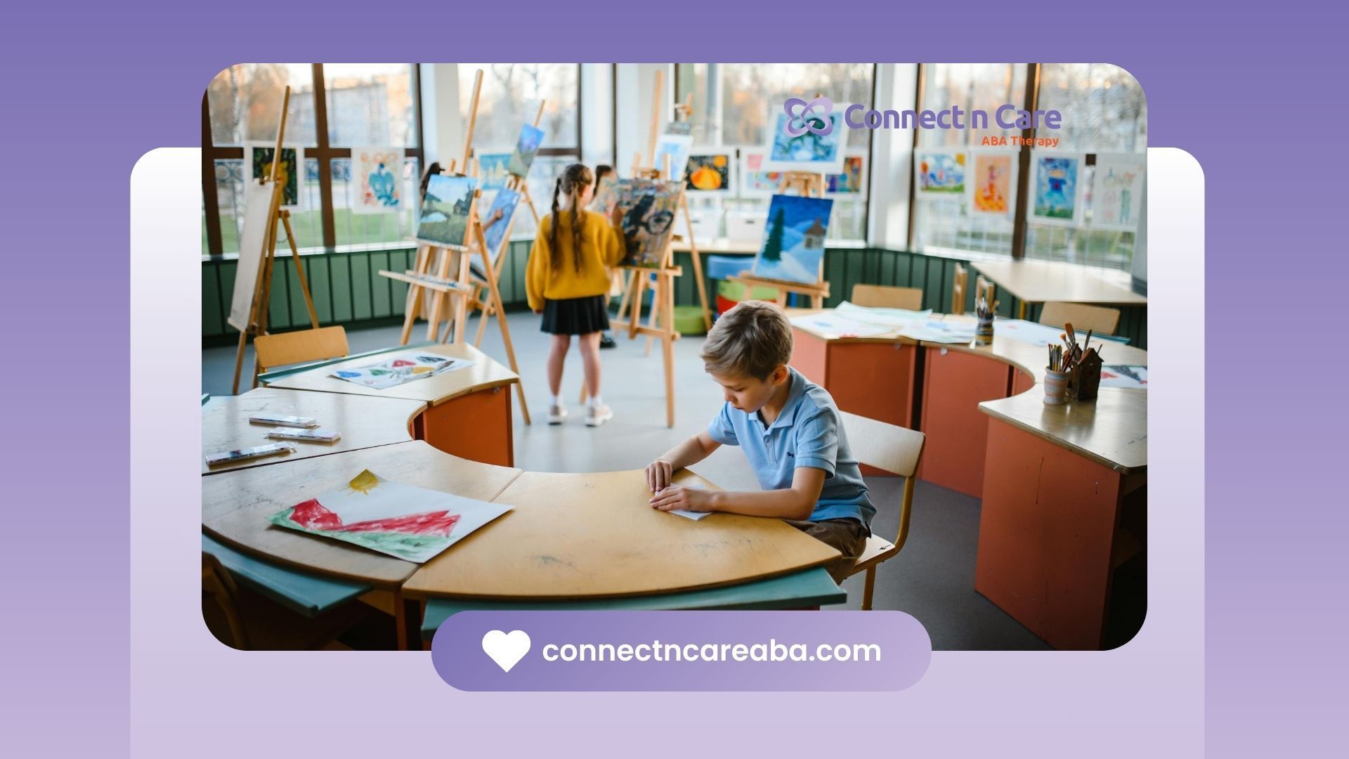 An autistic child sitting at a desk in an art classroom with colorful paintings displayed on easels.