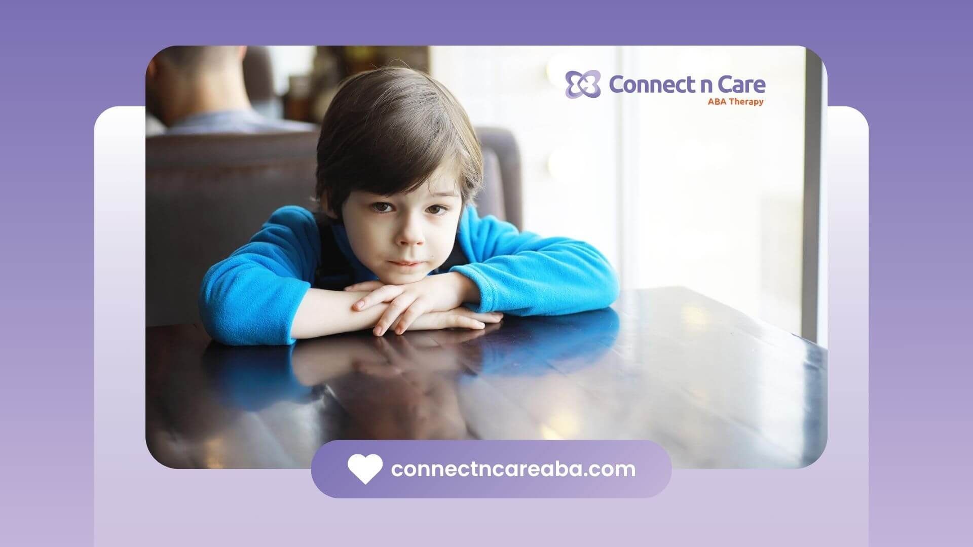 A young boy with autism in a blue sweater is leaning on a table.