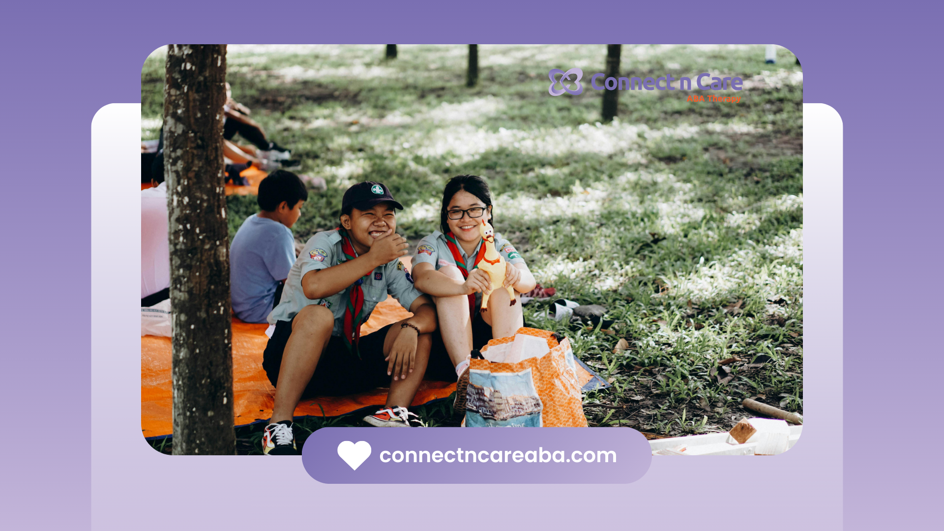Two people smile while sitting on an orange blanket under trees. One person has a hat and the other wears glasses.