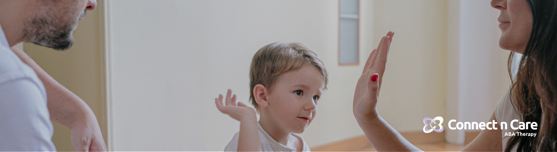A family, with a toddler, interacts in a room with plain walls. The mother high-fives the child.