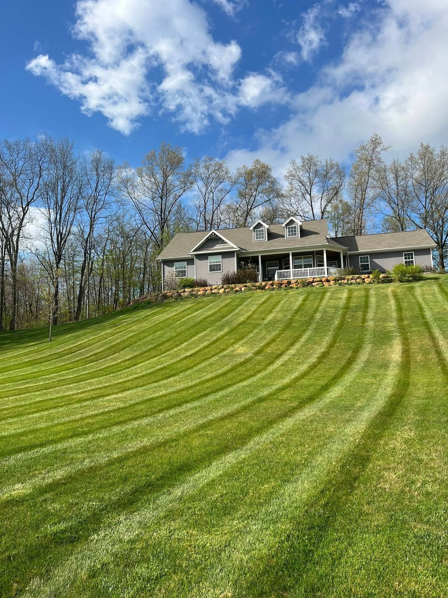 A house sits atop a hill with a mown lawn featuring distinctive dark and light green grass stripes under a blue sky.