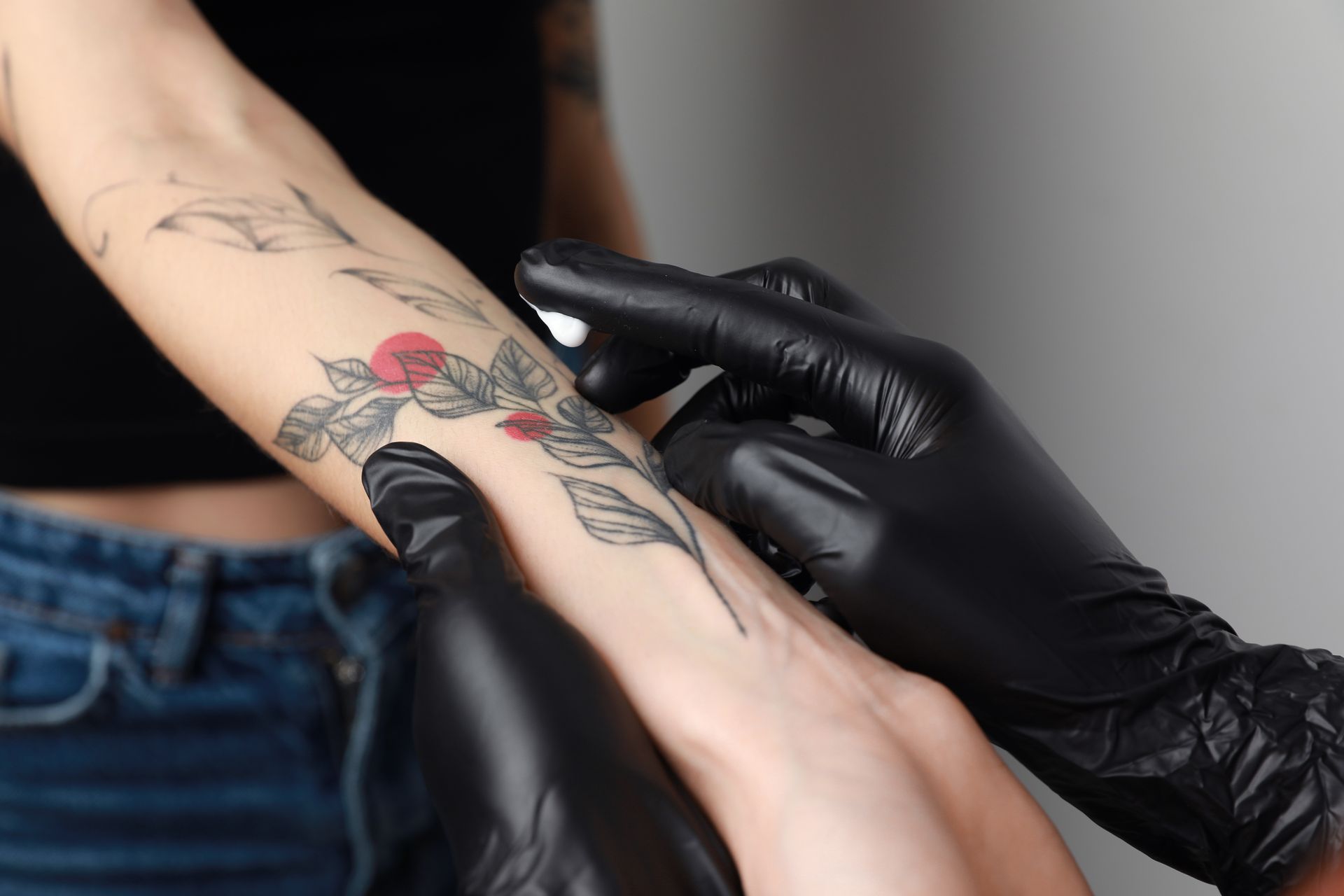 Worker in gloves applying cream on woman's arm with tattoo against light background, closeup.