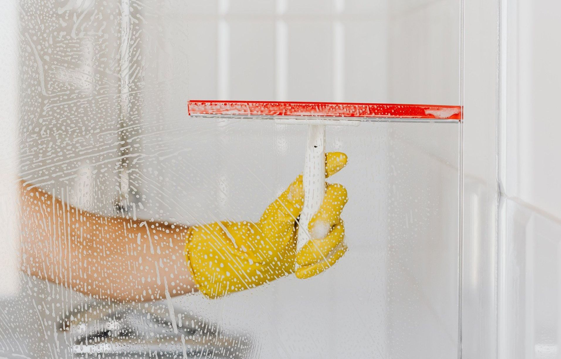 A person wearing yellow gloves is cleaning a shower door with a squeegee.
