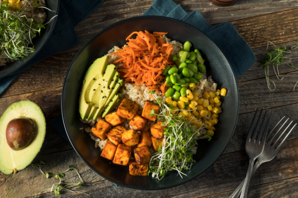 A colorful Buddha bowl with avocado, tofu, carrots, edamame, corn, sprouts, and rice, on a wooden table.