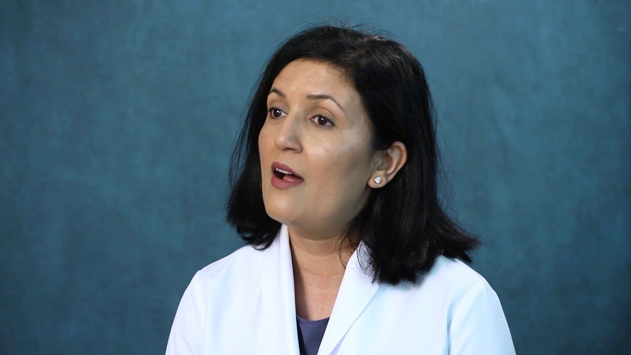 Woman with dark hair and a white coat speaks against a blue background.