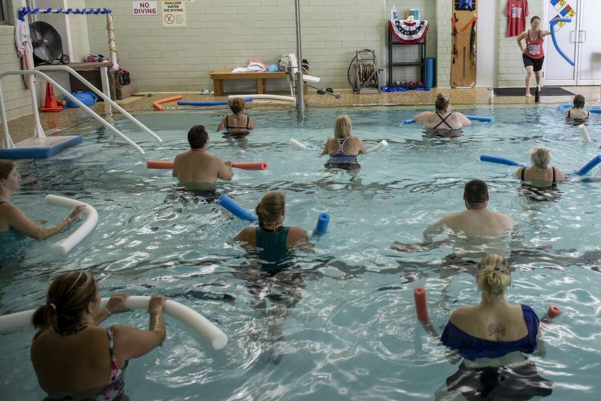 People doing water aerobics in a pool, using foam noodles for exercise. The instructor stands near the door.