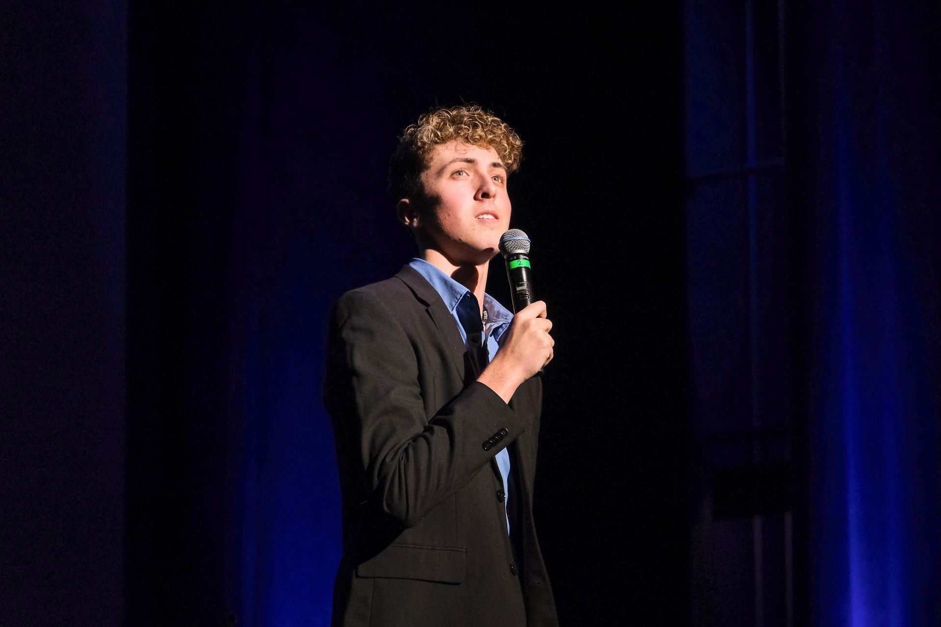 Young person in a suit, holding a microphone, speaking on stage with blue lighting.