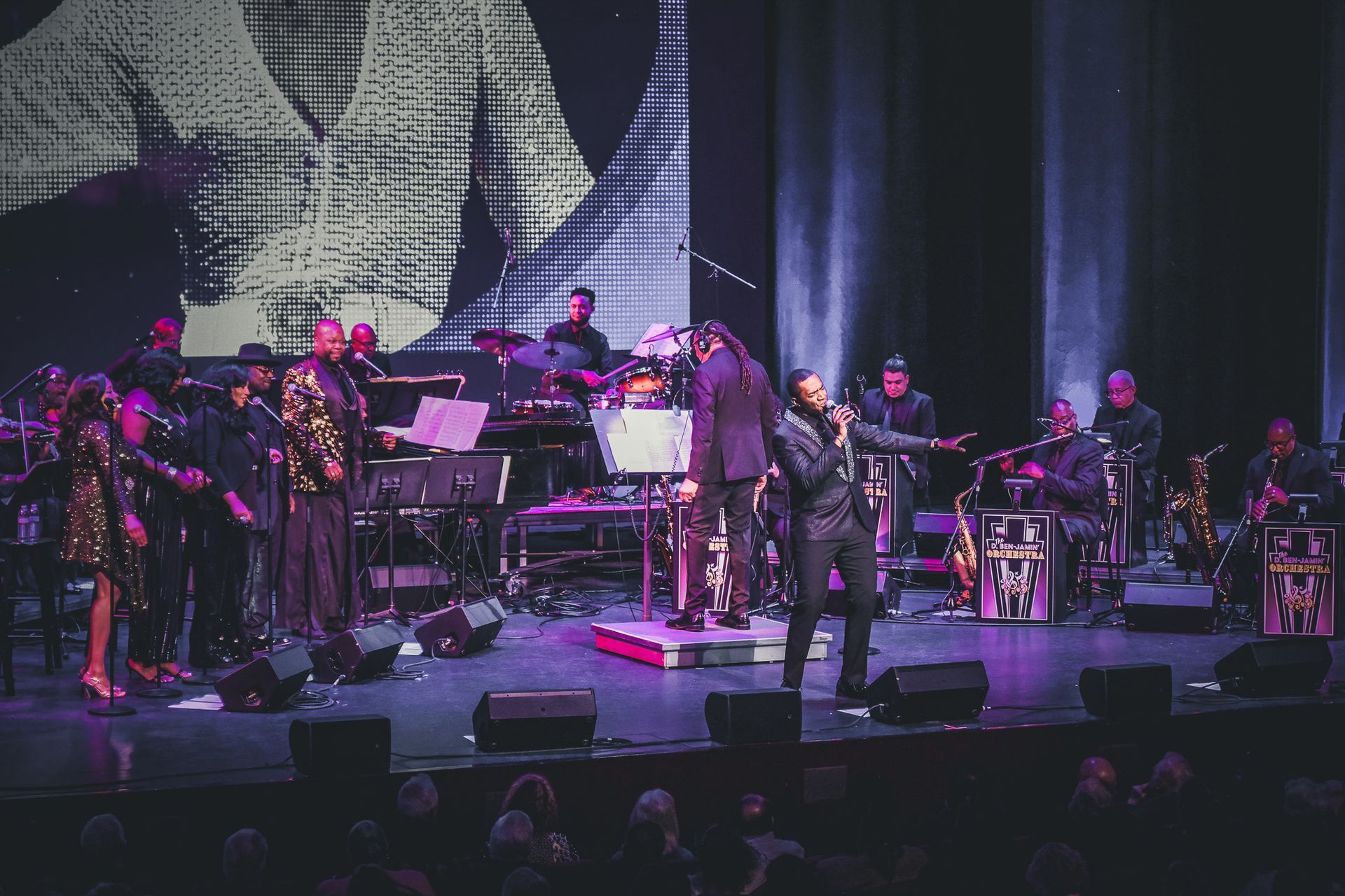A musical performance with a band on stage, singer in focus. Purple stage lighting, large screen backdrop.