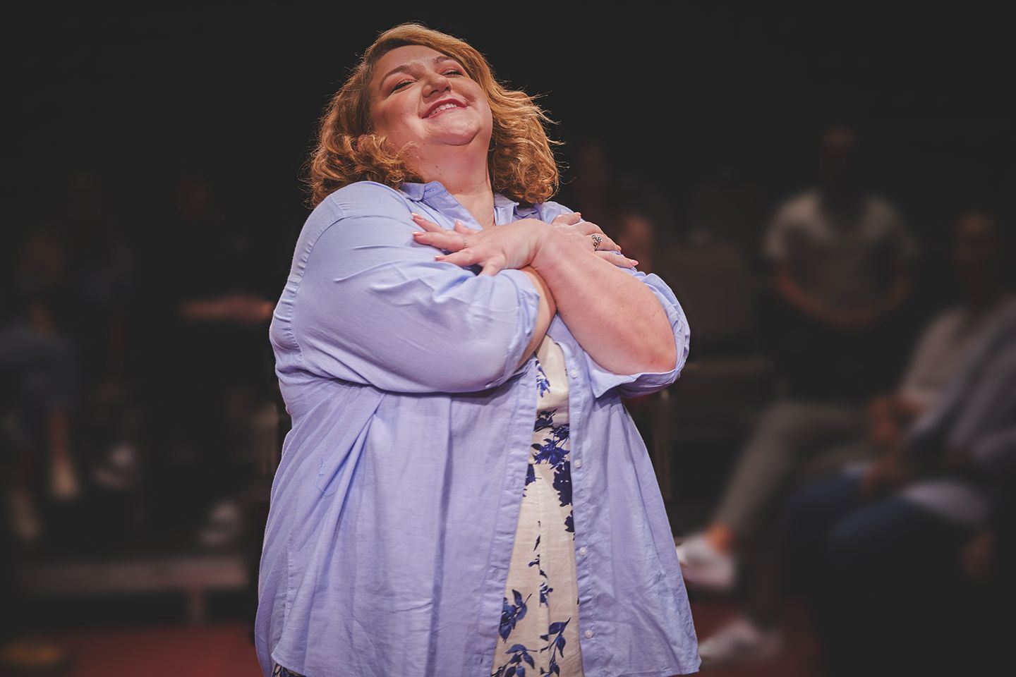 Woman on stage with arms crossed, smiling. Wearing a light blue shirt over a floral dress. Dark background, blurred audience.