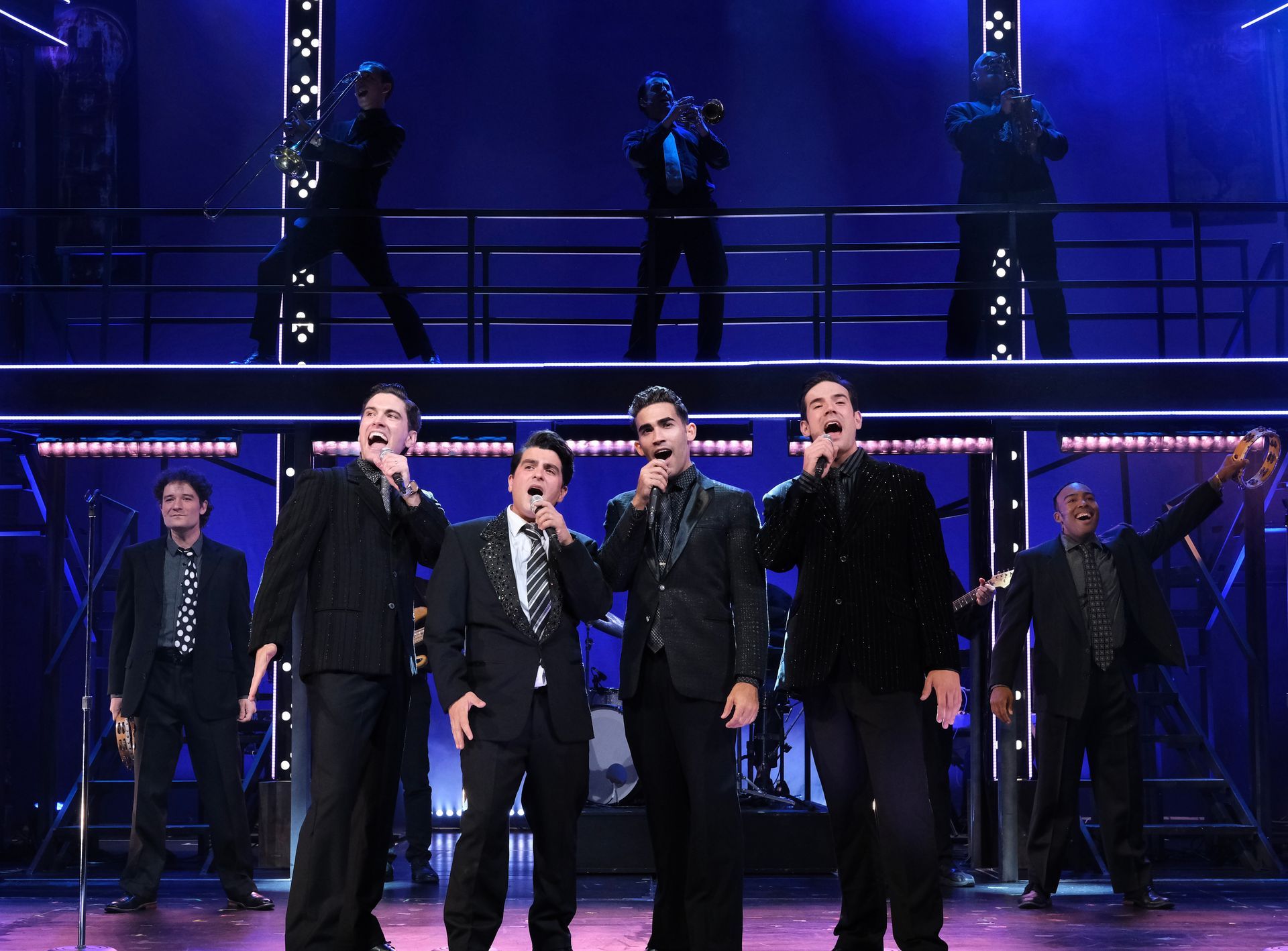 Men in black suits singing on stage with a band in the background. Bright lights.