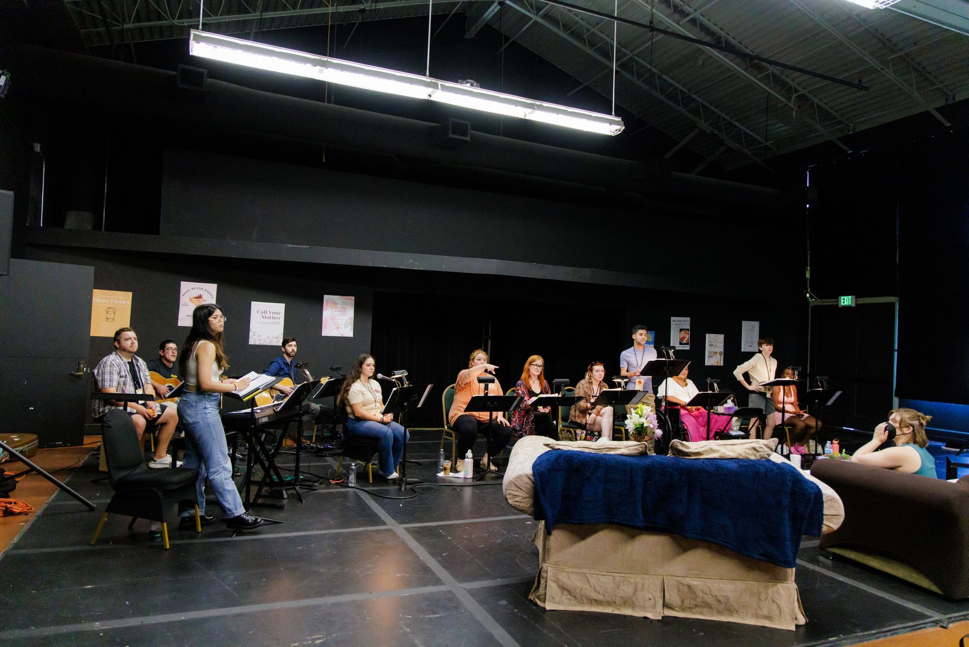 A group of people rehearse music in a dimly lit studio with black walls and a stage.