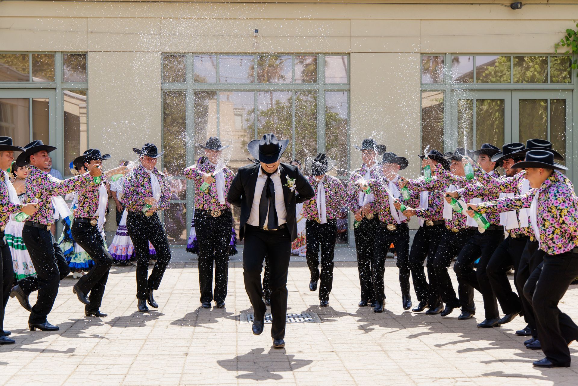 Dancers in black hats and colorful shirts throw confetti, celebrating in front of a building with large windows.