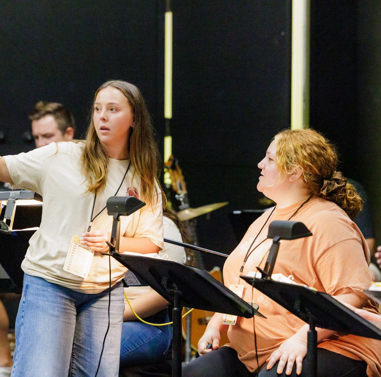 Two women in an orchestra setting; one pointing and the other looking on, both wearing lanyards and near music stands.