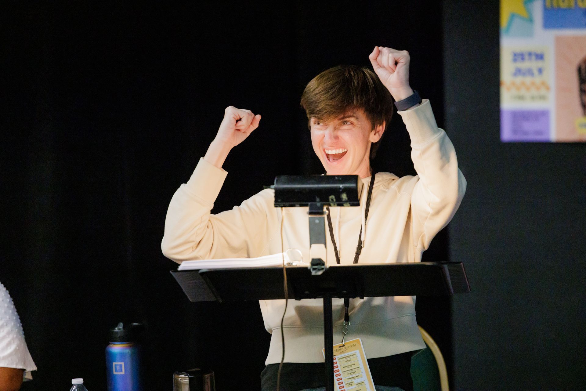 Person with raised fists and joyful expression behind a lectern; indoor setting with black backdrop and poster.