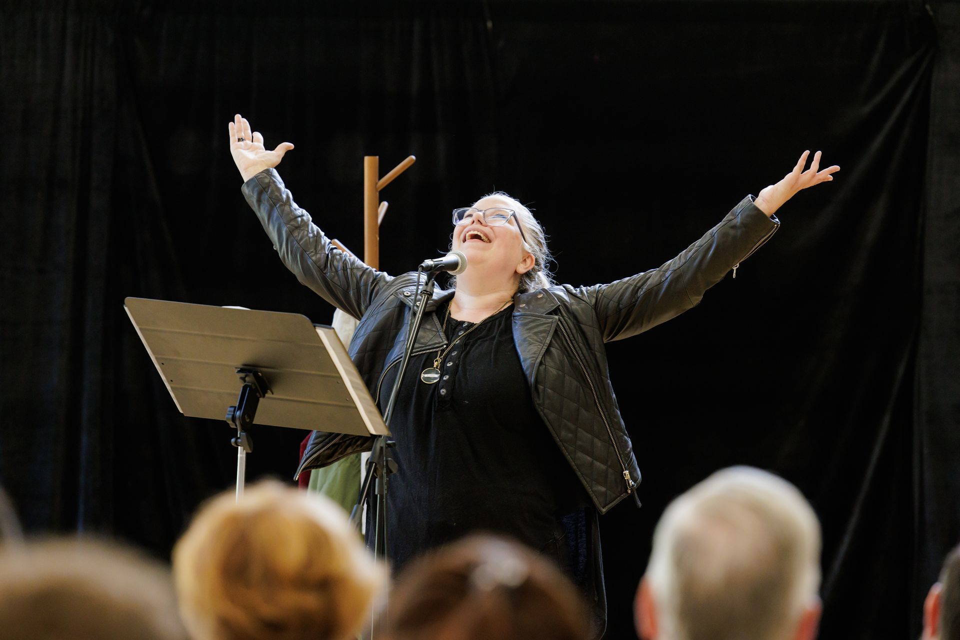 Woman on stage with arms raised, singing into a microphone. Dark background, audience in foreground.