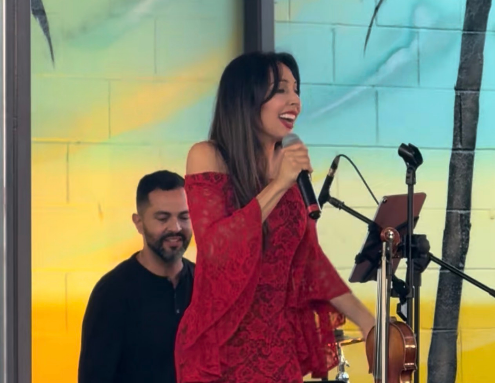 Woman in red dress sings into a microphone, man in black shirt smiles in the background, by a wall with mural.