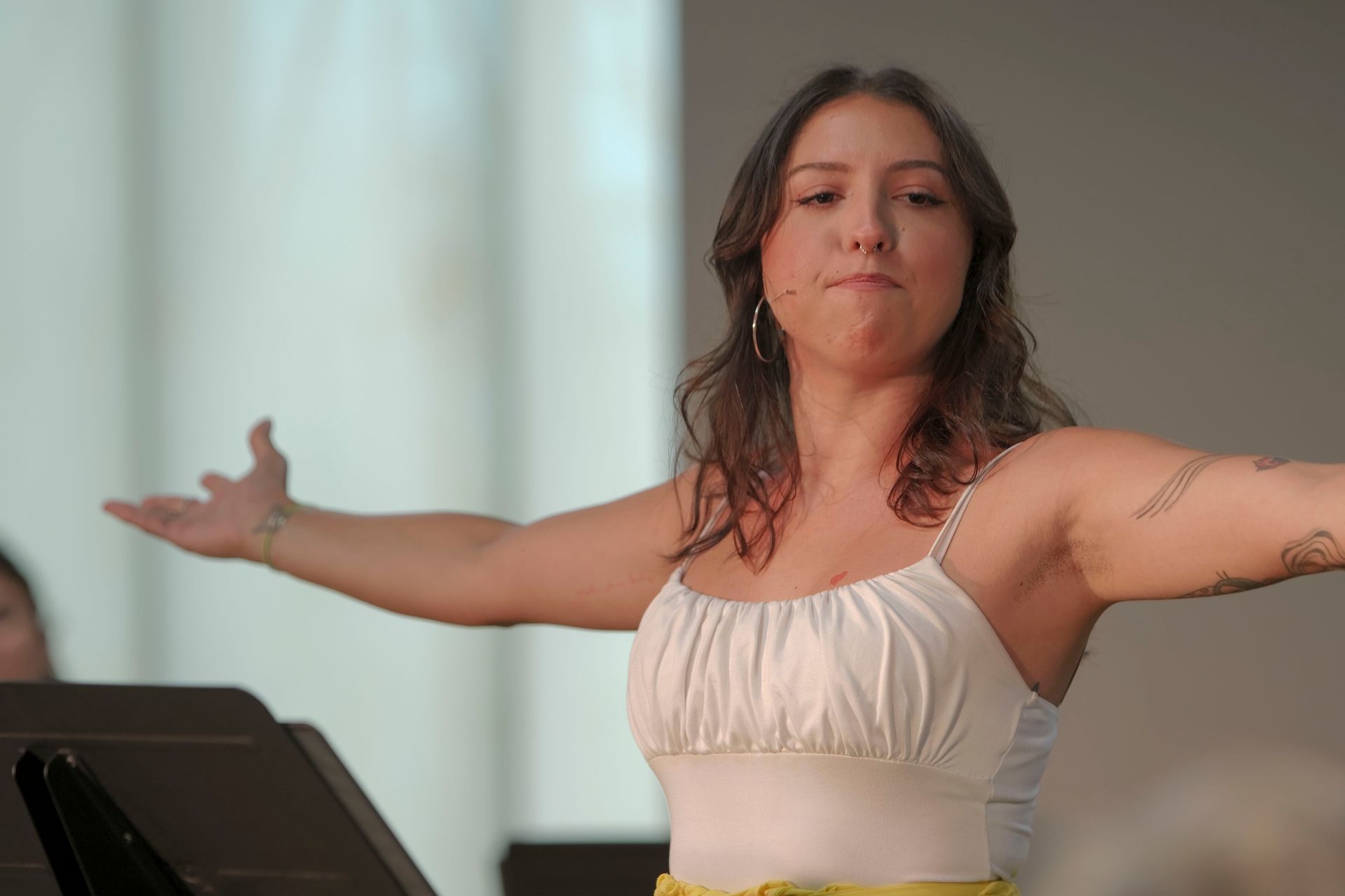 Woman with arms outstretched, singing. White top, yellow skirt. Stage setting, music stand in foreground.