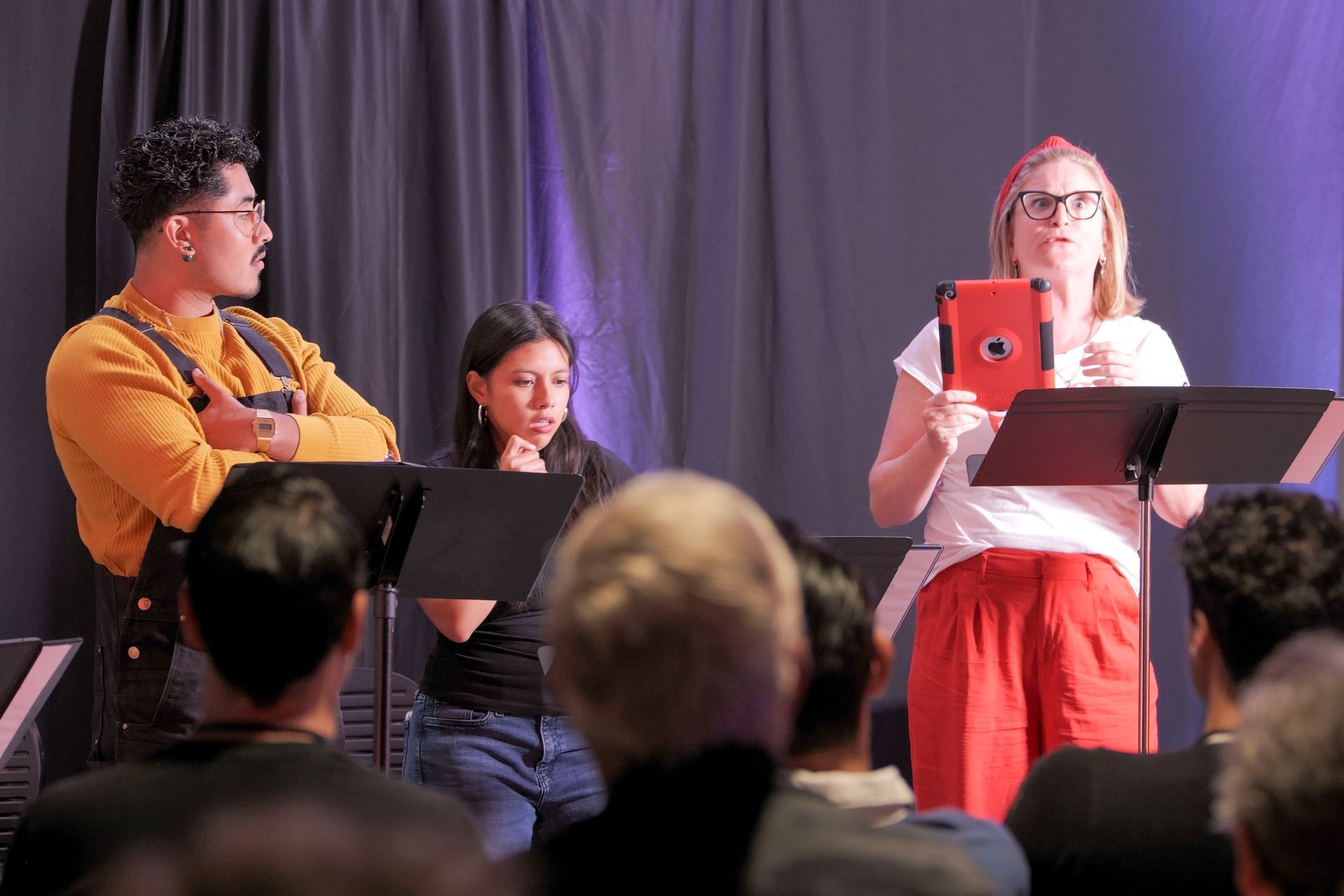 Three people performing on stage, reading from scripts. The woman in red pants holds a tablet.